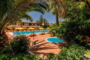 A pool area with multiple sun loungers and umbrellas, surrounded by lush greenery and palm trees. - Cala Ginepro Hotel Resort (Orosei)