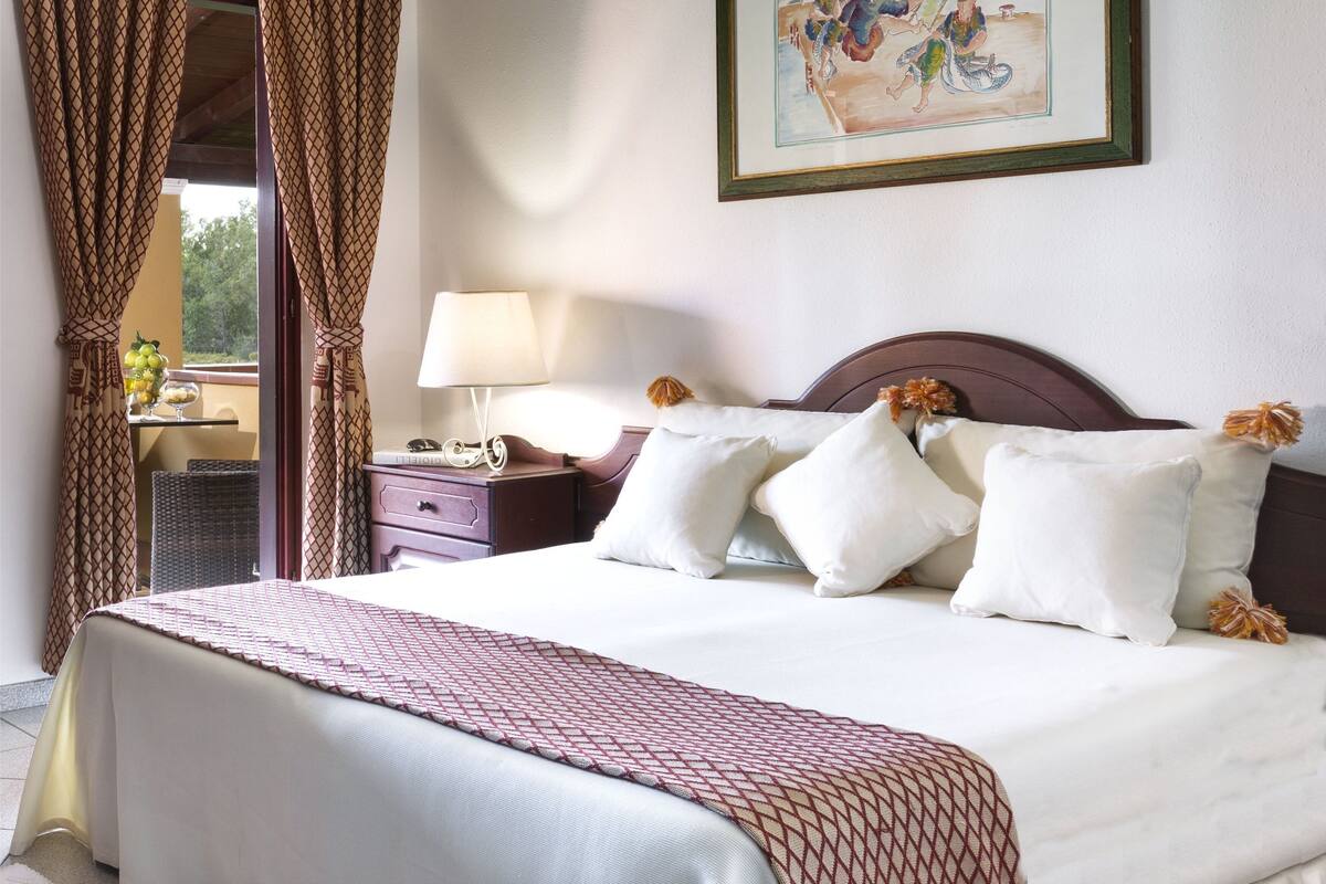A hotel room with a bed, a nightstand, a lamp, a framed picture on the wall, and a view of the outdoors through a window with curtains.