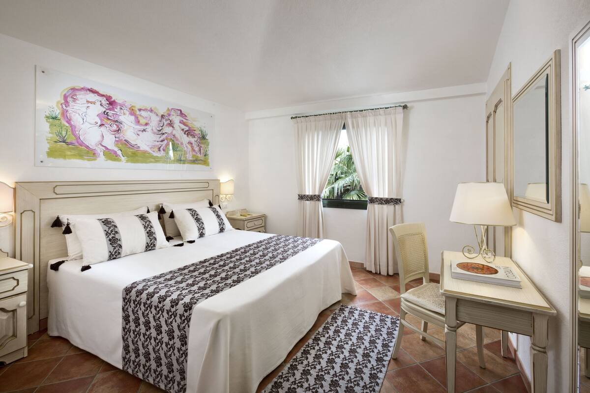 A bedroom with a large bed, a nightstand, a chair, a desk with a lamp, and a window with curtains.