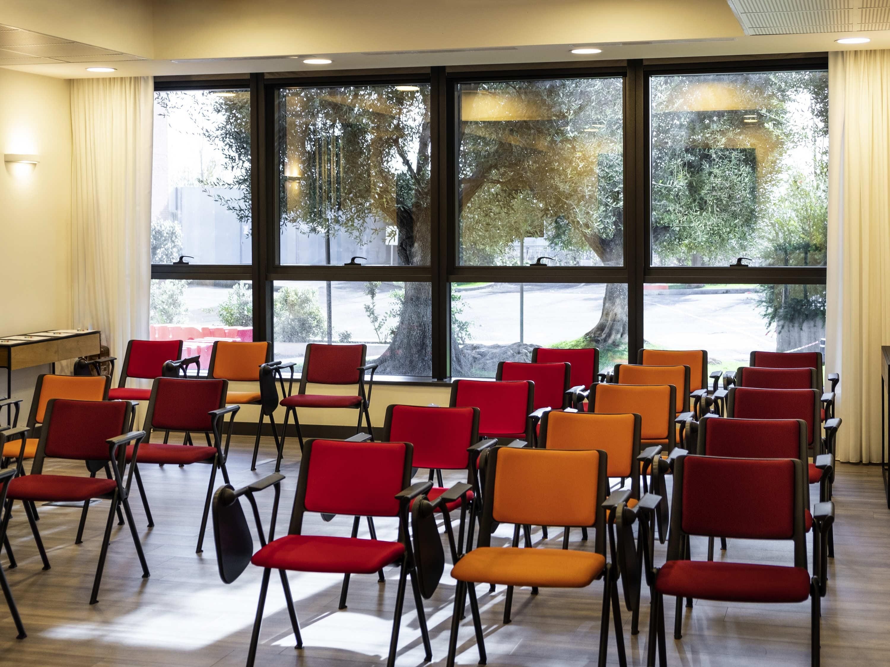 Una sala conferenze con file di sedie, ampie finestre che danno su alberi e un parcheggio, e un tavolo con una lavagna.