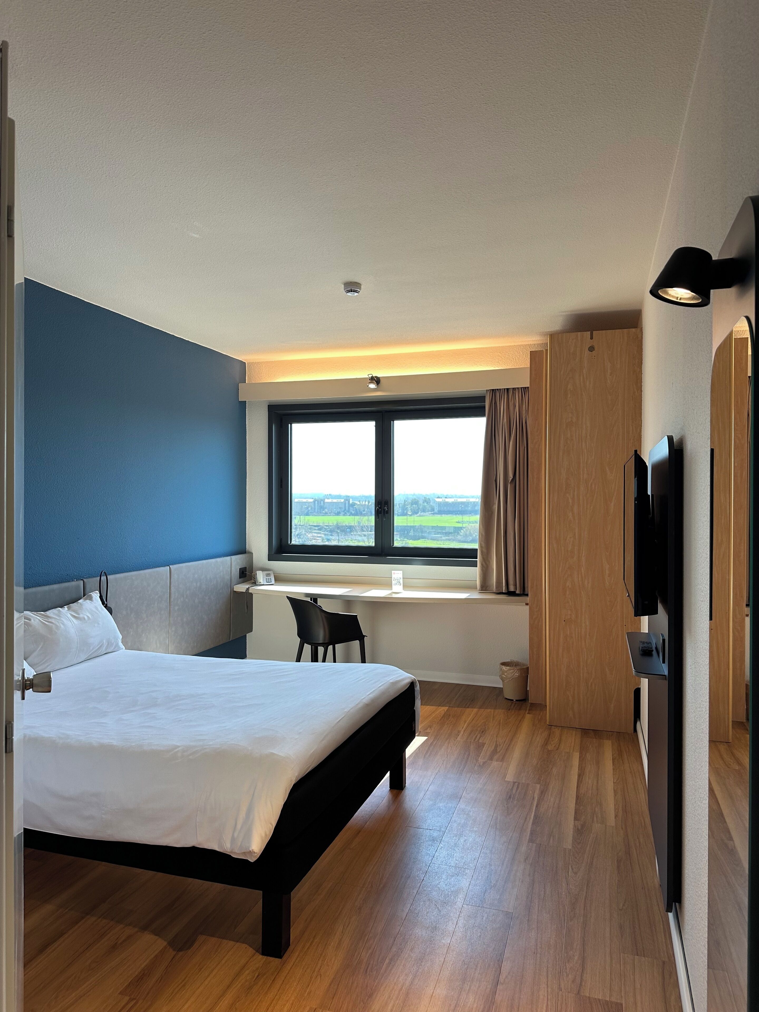 A hotel room with a bed, desk, chair, TV, and a large window with a view.