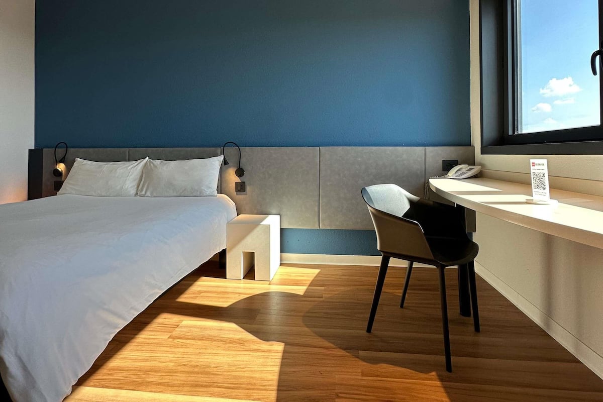 A modern hotel room with a bed, a desk, a chair, and a window with a view of the sky.