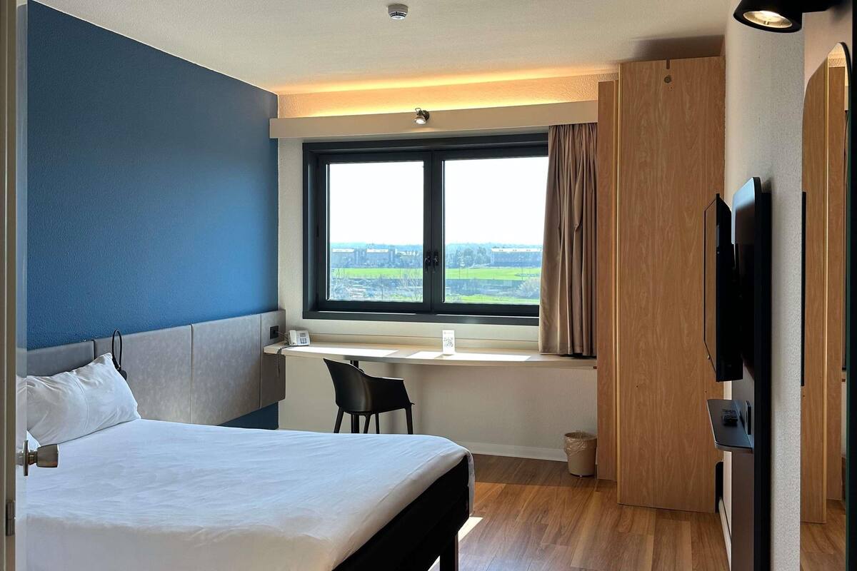 A hotel room with a bed, a desk, a chair, a TV, and a window with a view.