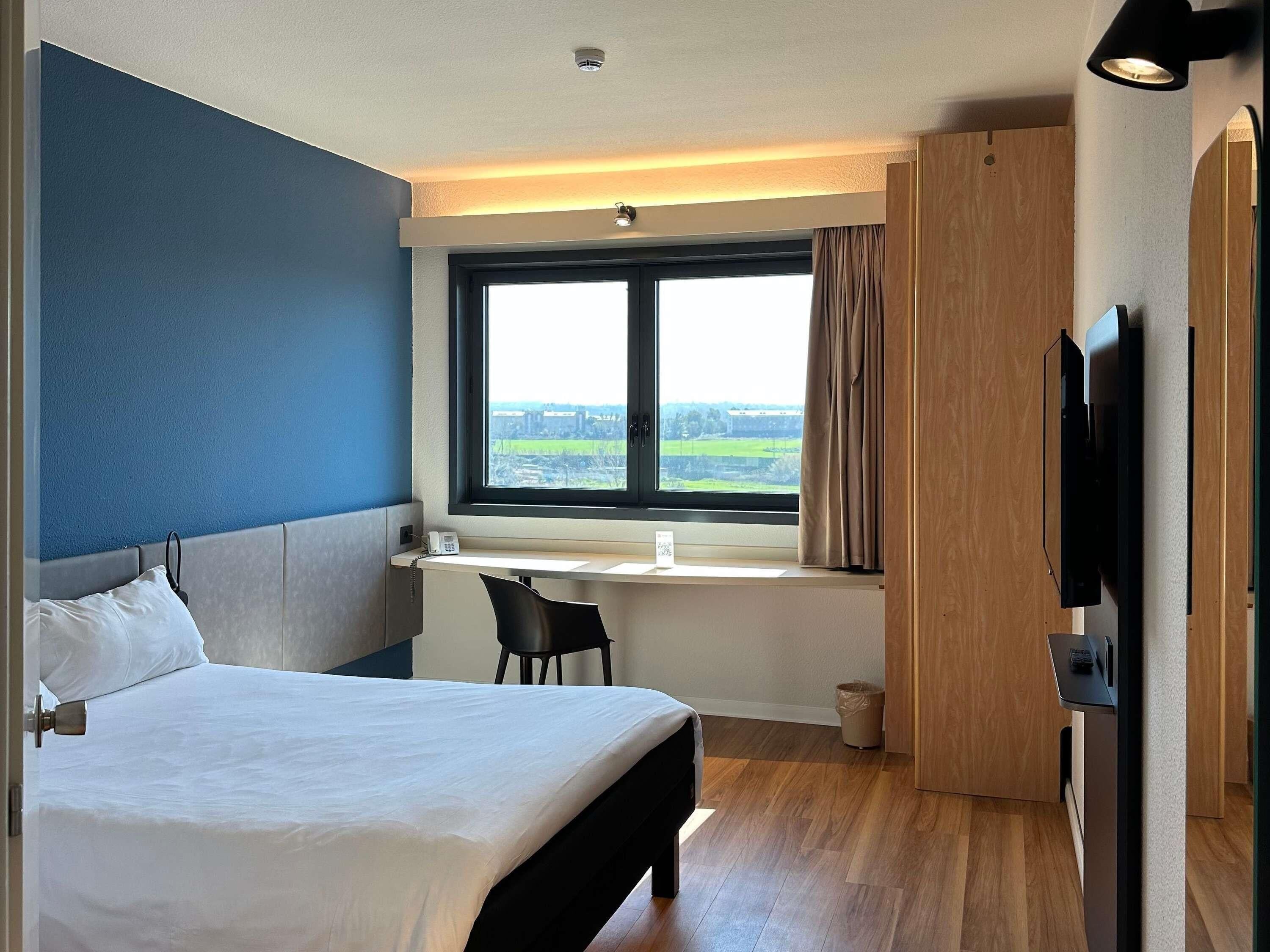 A hotel room with a bed, a desk, a chair, a TV, and a window with a view.