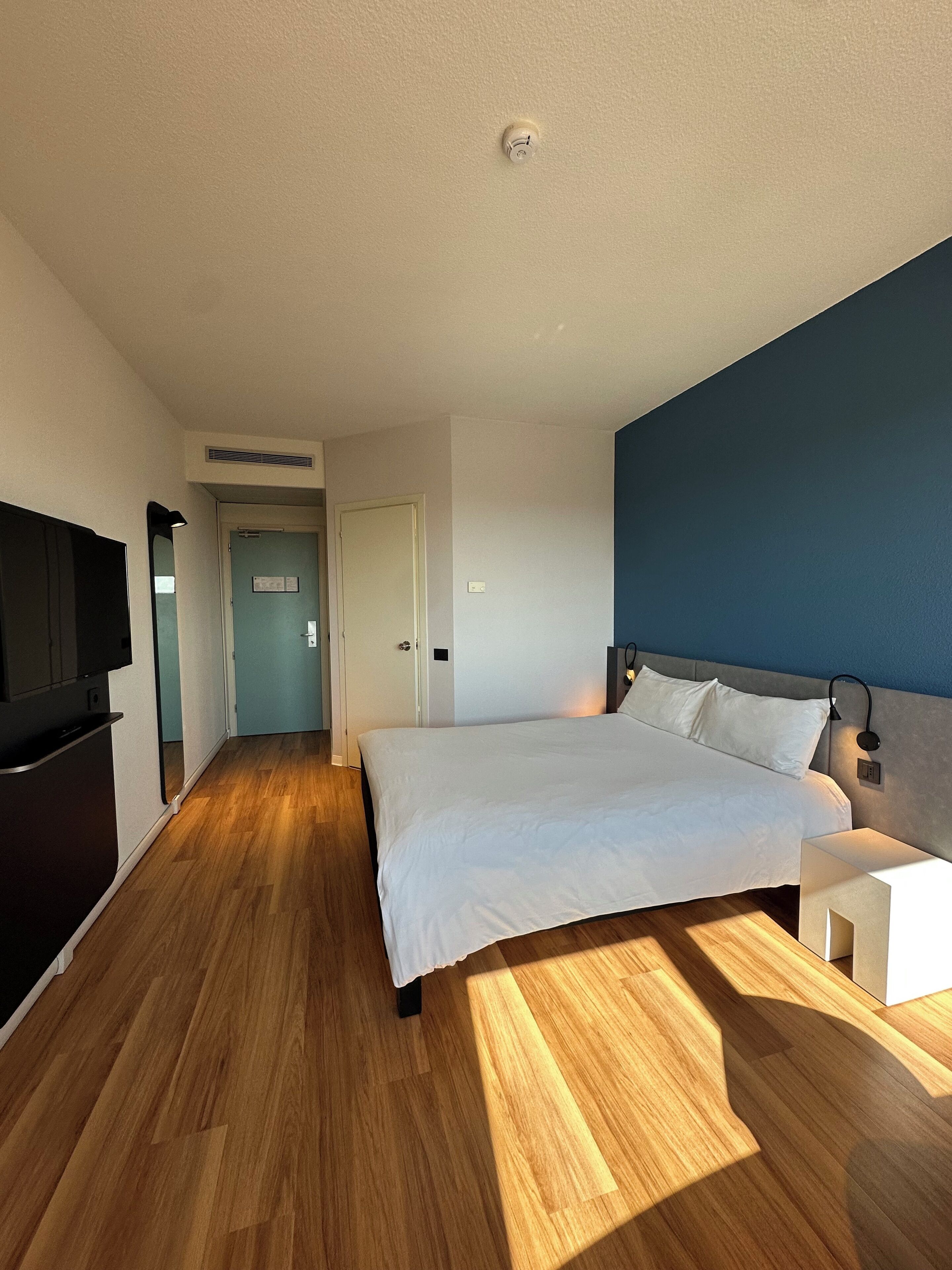 A modern hotel room with a wooden floor, a large bed, a TV, and a blue accent wall.