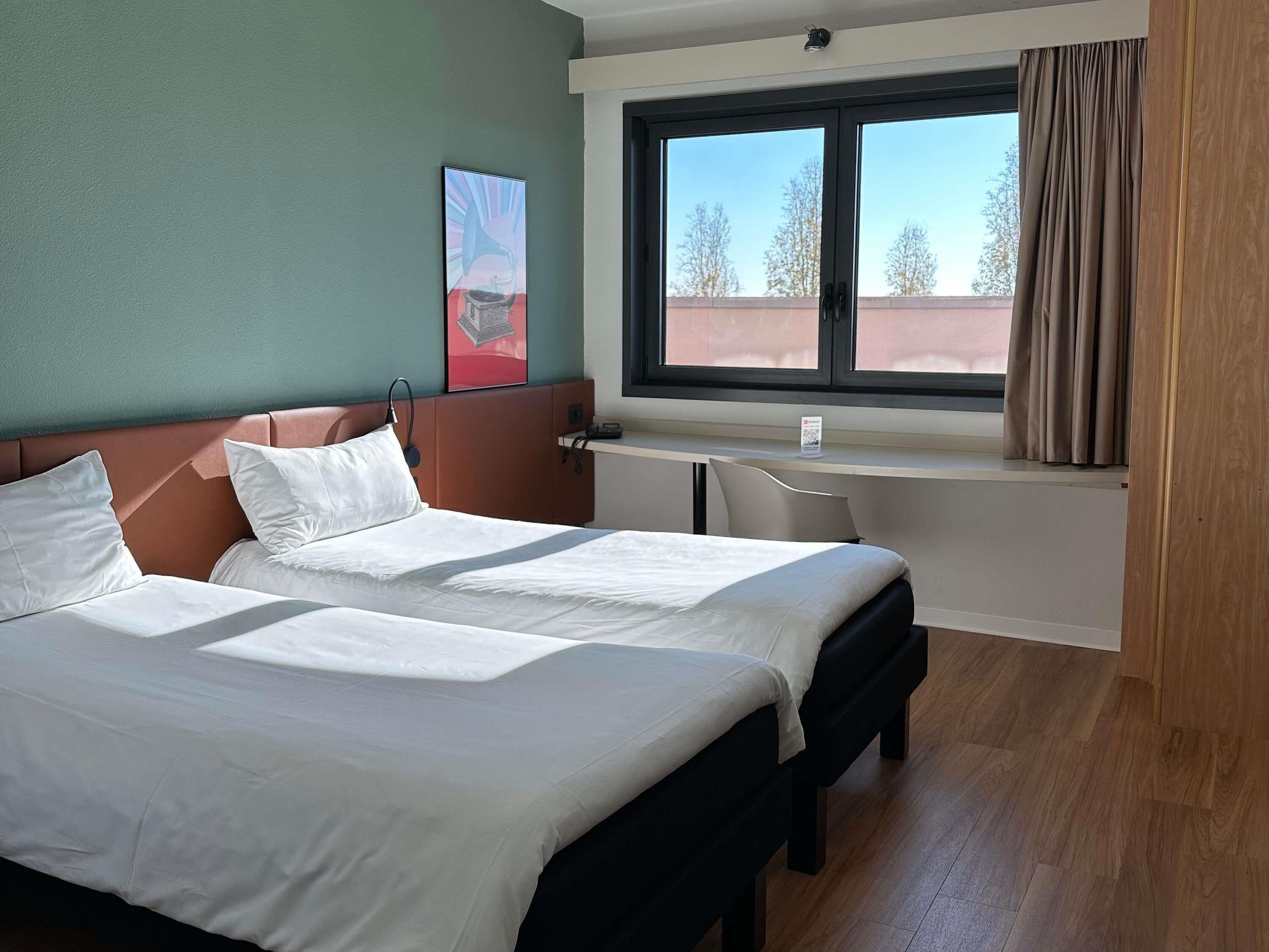 A hotel room with a bed, a desk, a window with curtains, and a painting on the wall.