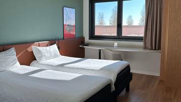 A hotel room with a bed, a desk, a window with curtains, and a painting on the wall.