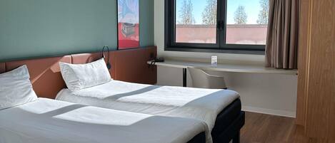Et hotelværelse med en seng, et skrivebord, et vindue med gardiner og et billede på væggen.