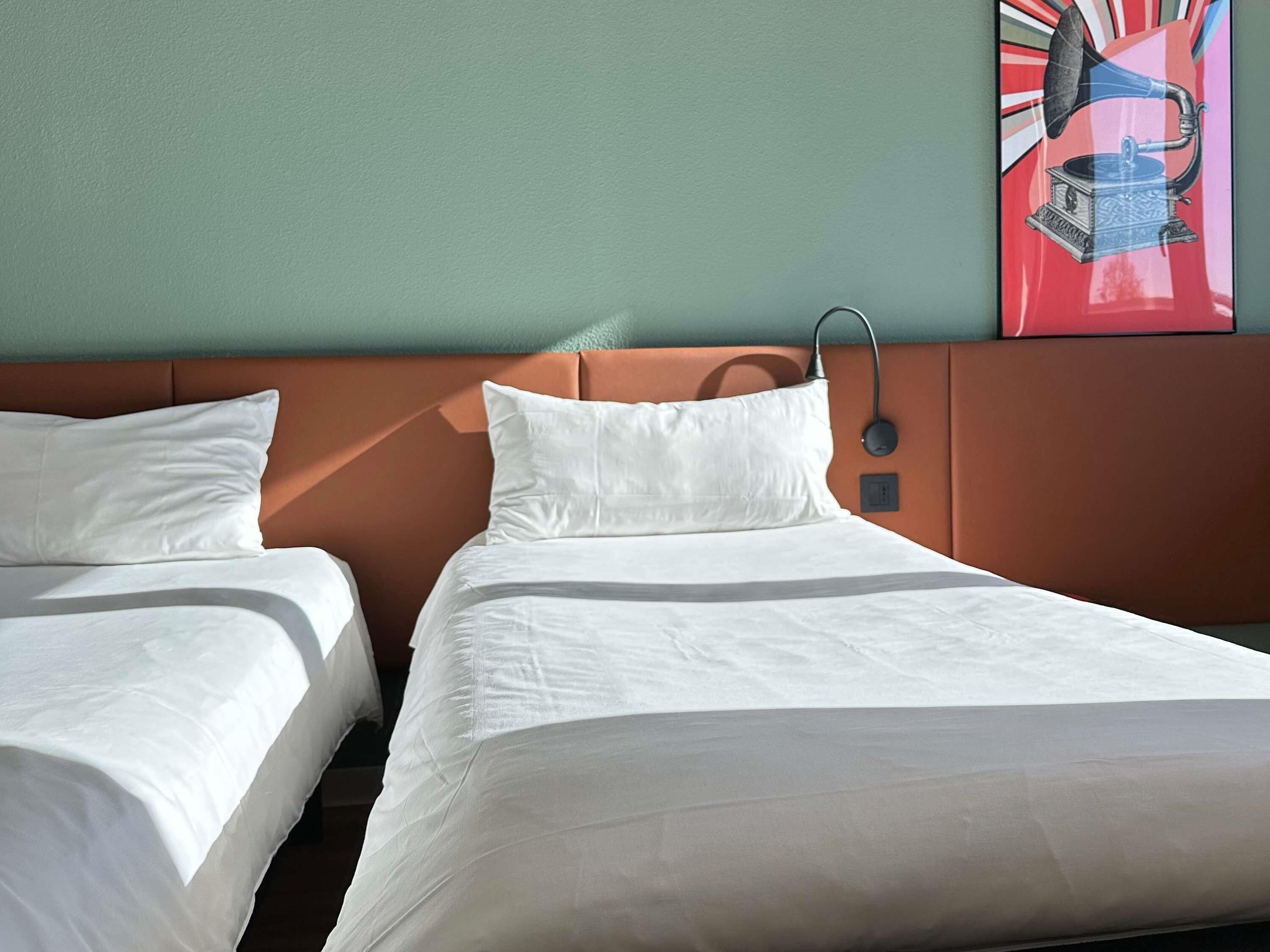 Quarto de hotel com duas camas de solteiro, cabeceira e luminária fixada na parede.