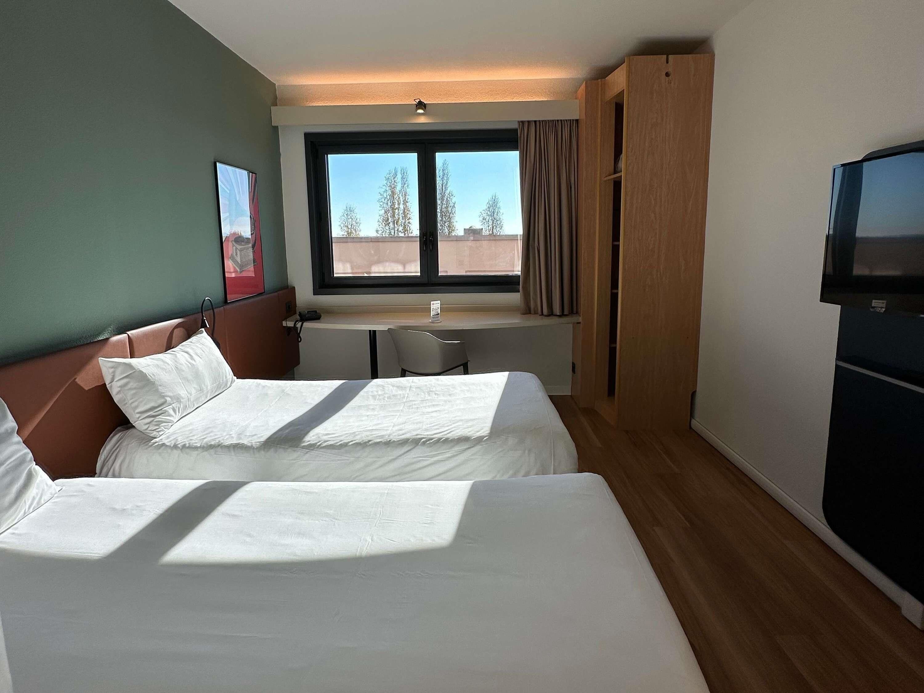 A hotel room with two beds, a desk, a TV, and a window with curtains.