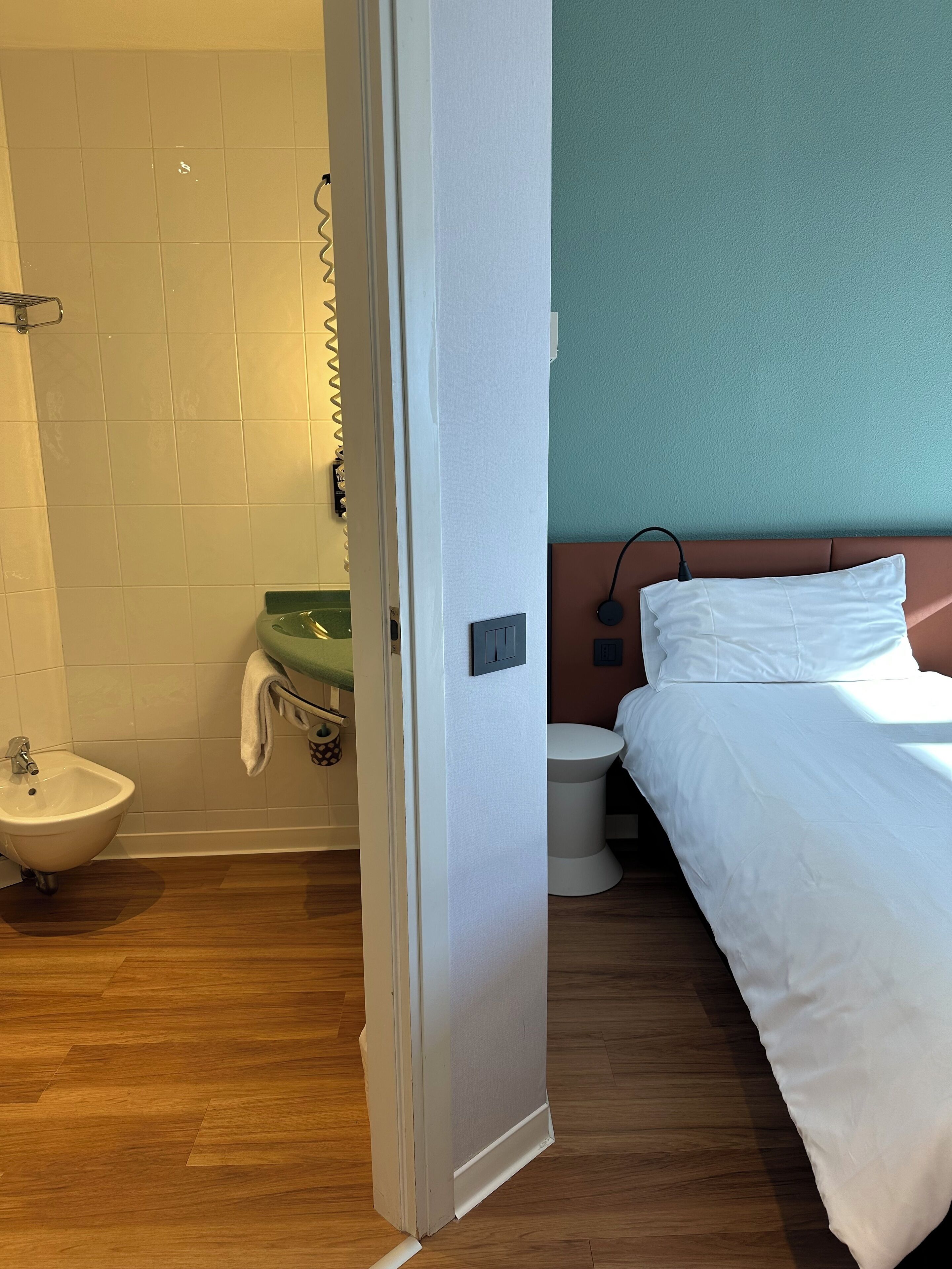 A hotel room with a wooden floor, a white bed, a green sink, and a white toilet.