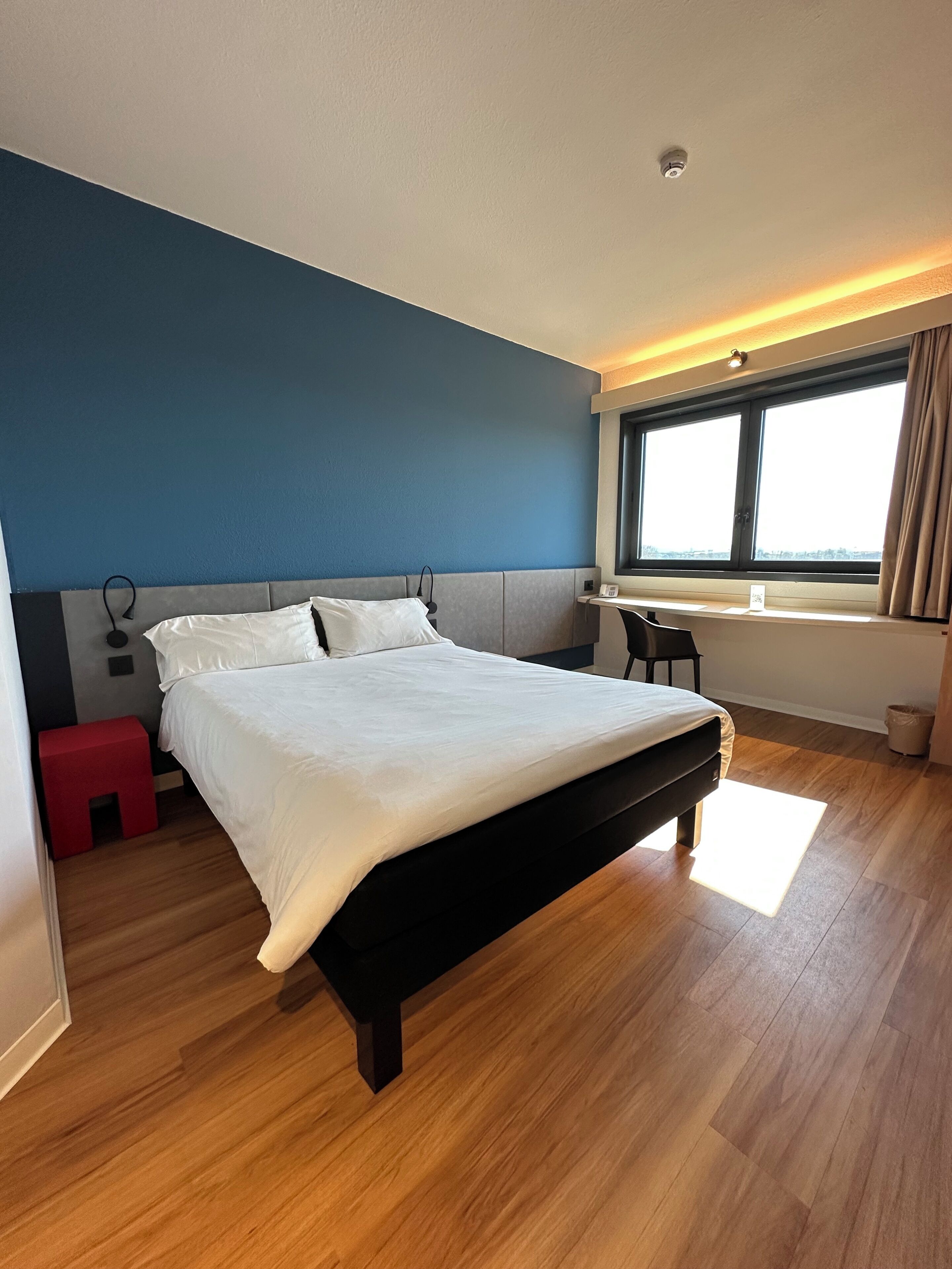 A modern hotel room with a large bed, a desk, a window with curtains, and a red stool.