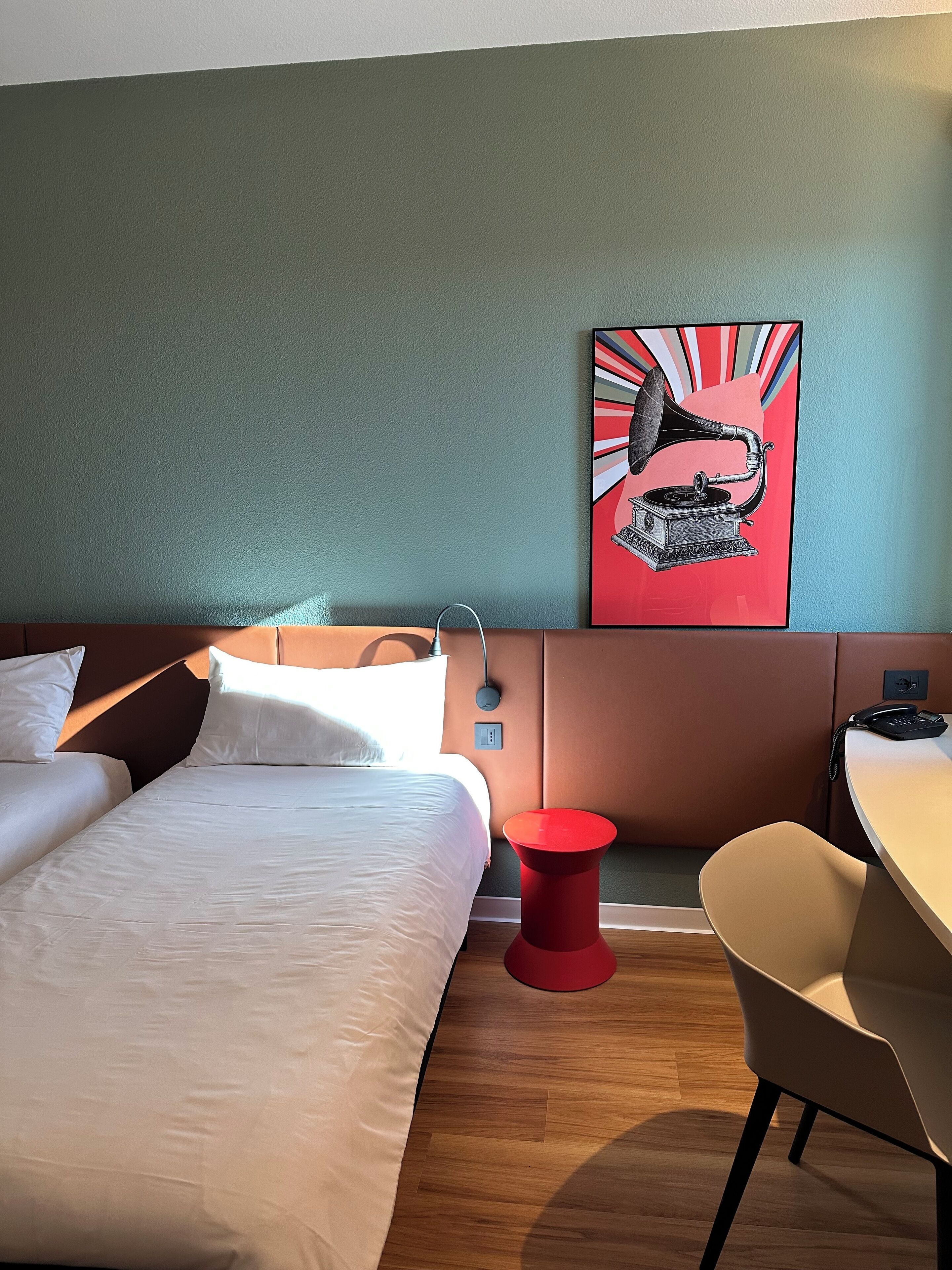 Habitación de hotel con cama, escritorio, silla, taburete rojo y un cuadro colgado en la pared de un fonógrafo.