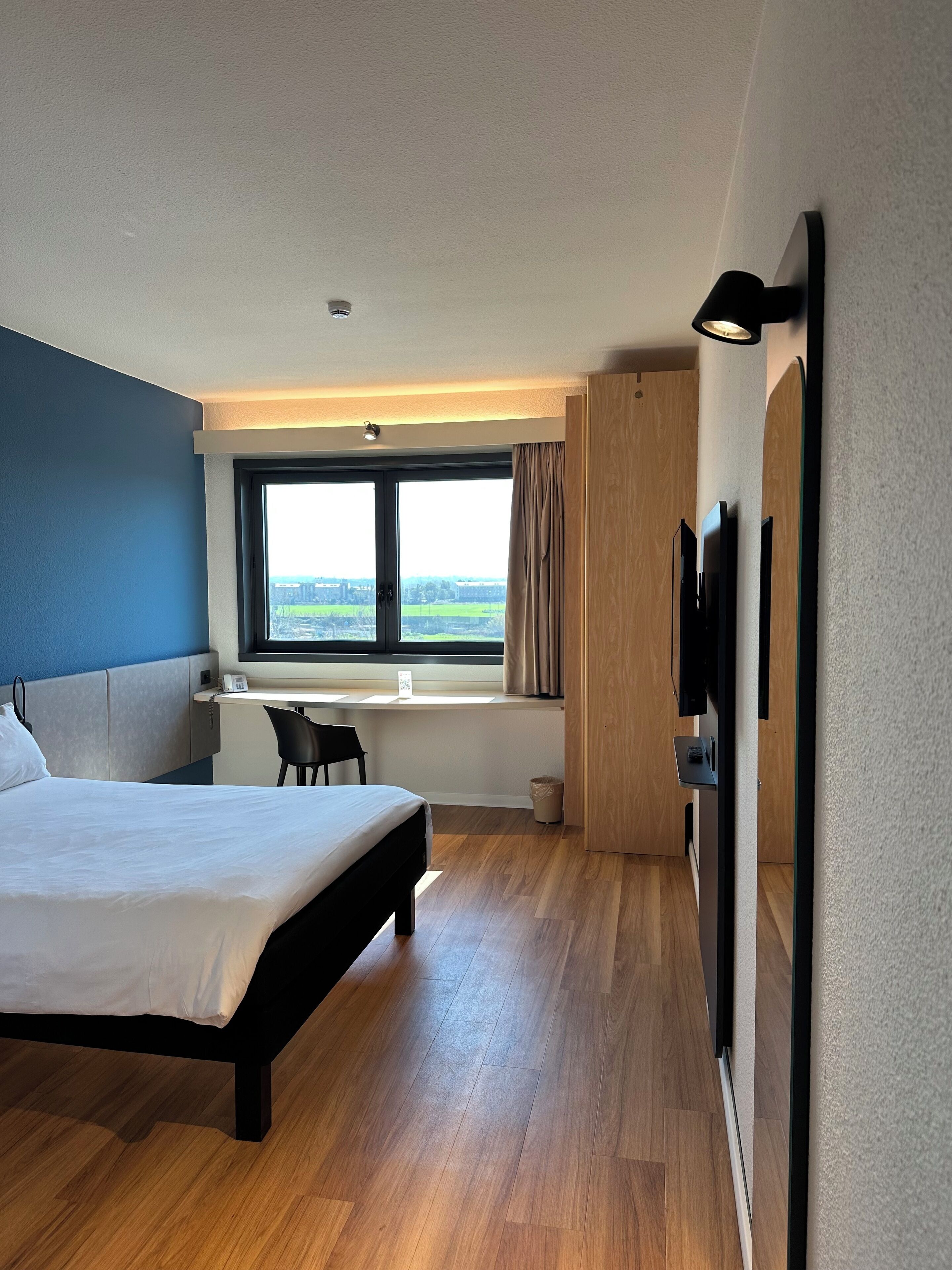 A modern hotel room with a large bed, a desk with a chair, a TV, and a window with a view of the countryside.
