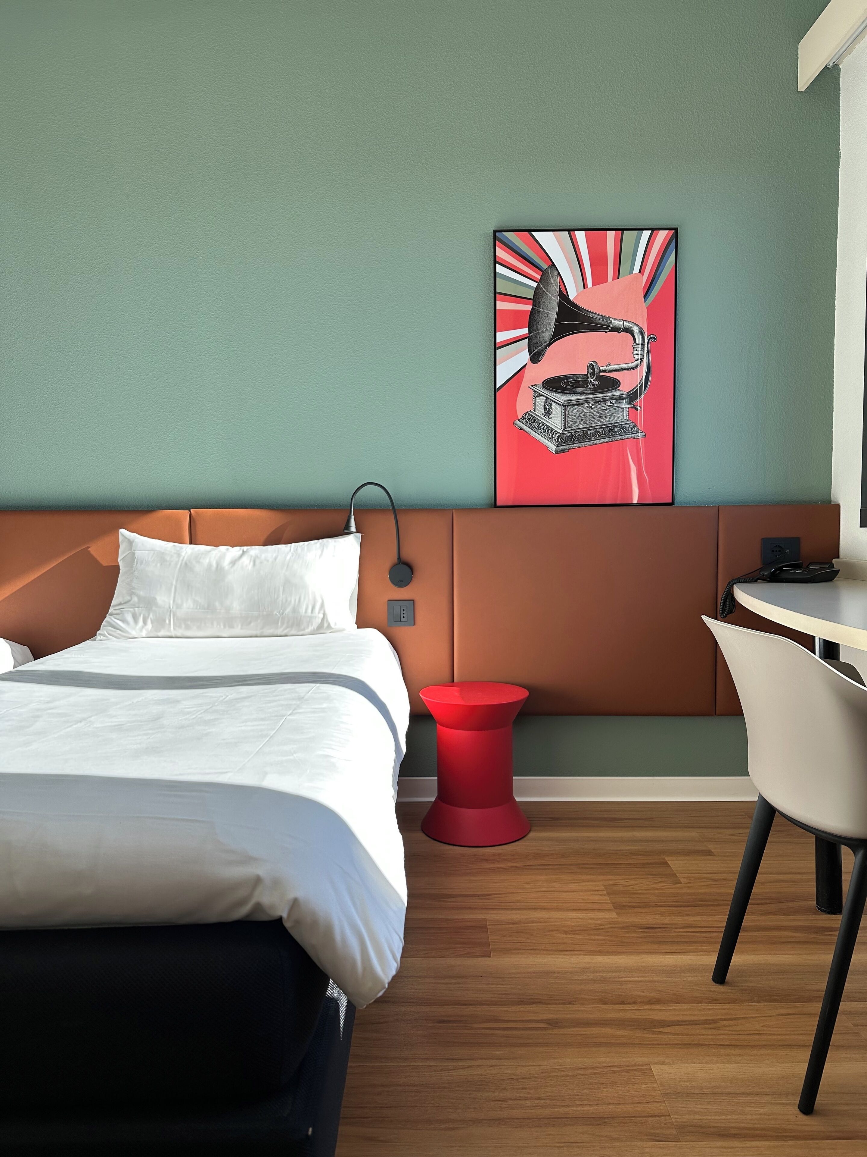 A hotel room with a bed, a red side table, a white chair, and a wall-mounted painting.