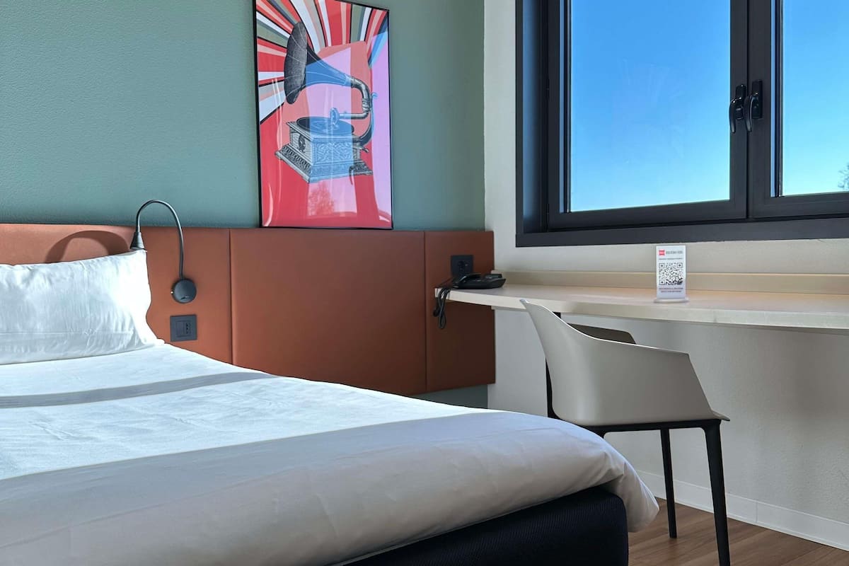 A hotel room with a bed, a desk, a chair, a window, and a wall art piece.