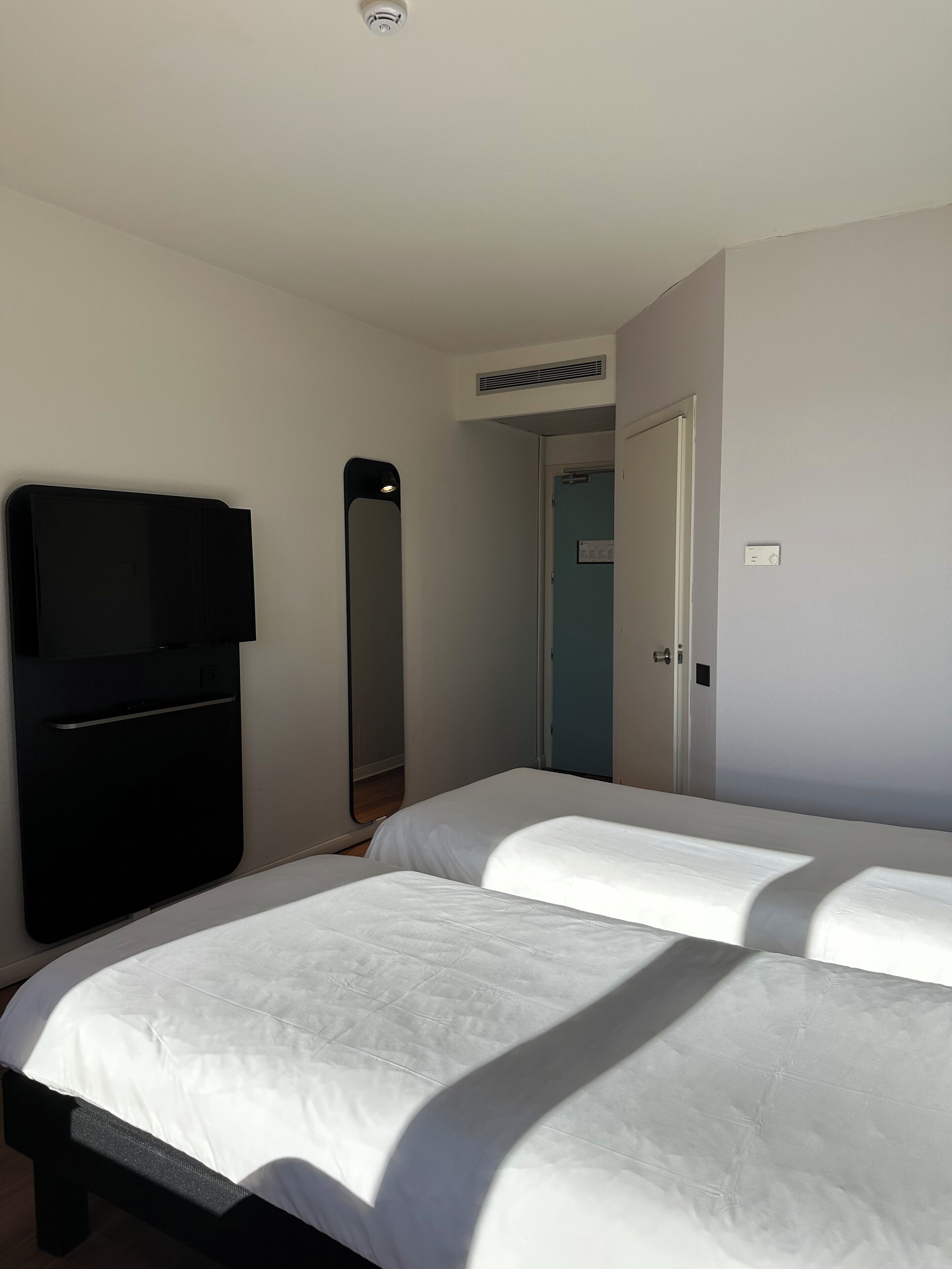 A hotel room with two beds, a flat-screen TV mounted on the wall, a door, and a light switch.