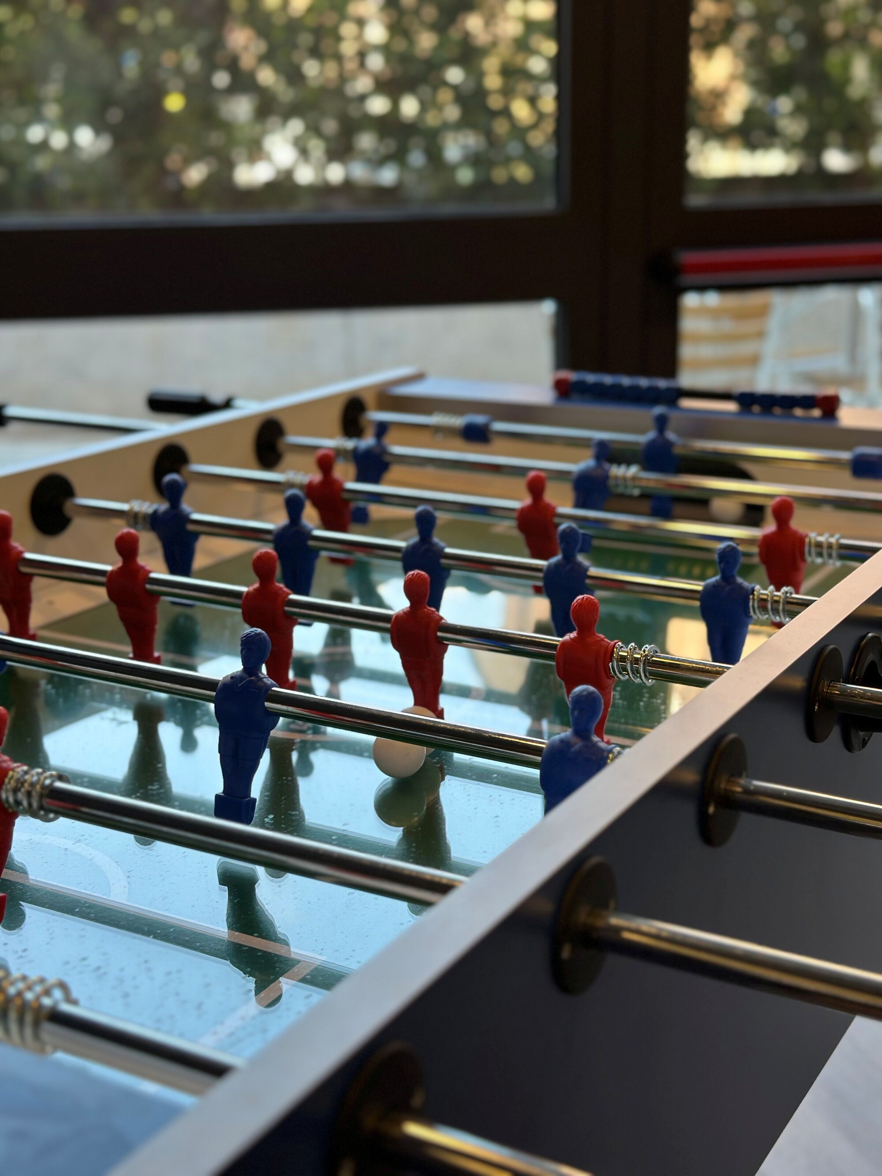 Una mesa de fútbol de mesa con figuras rojas y azules, frente a una ventana con vistas a árboles.