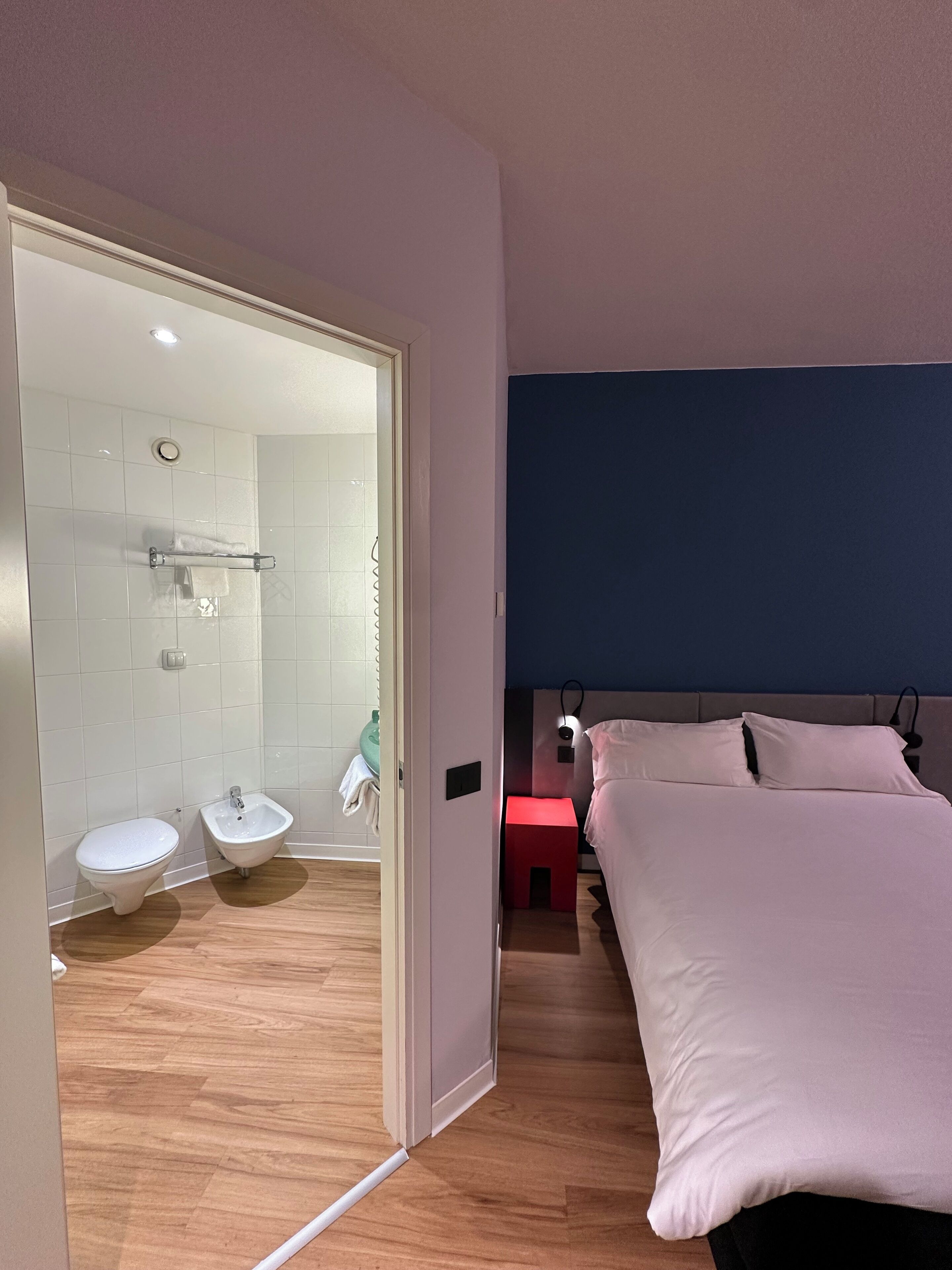 A modern bedroom with a bed, a nightstand, and a red stool. Adjacent is a bathroom with a bidet, a toilet, and a shower area.