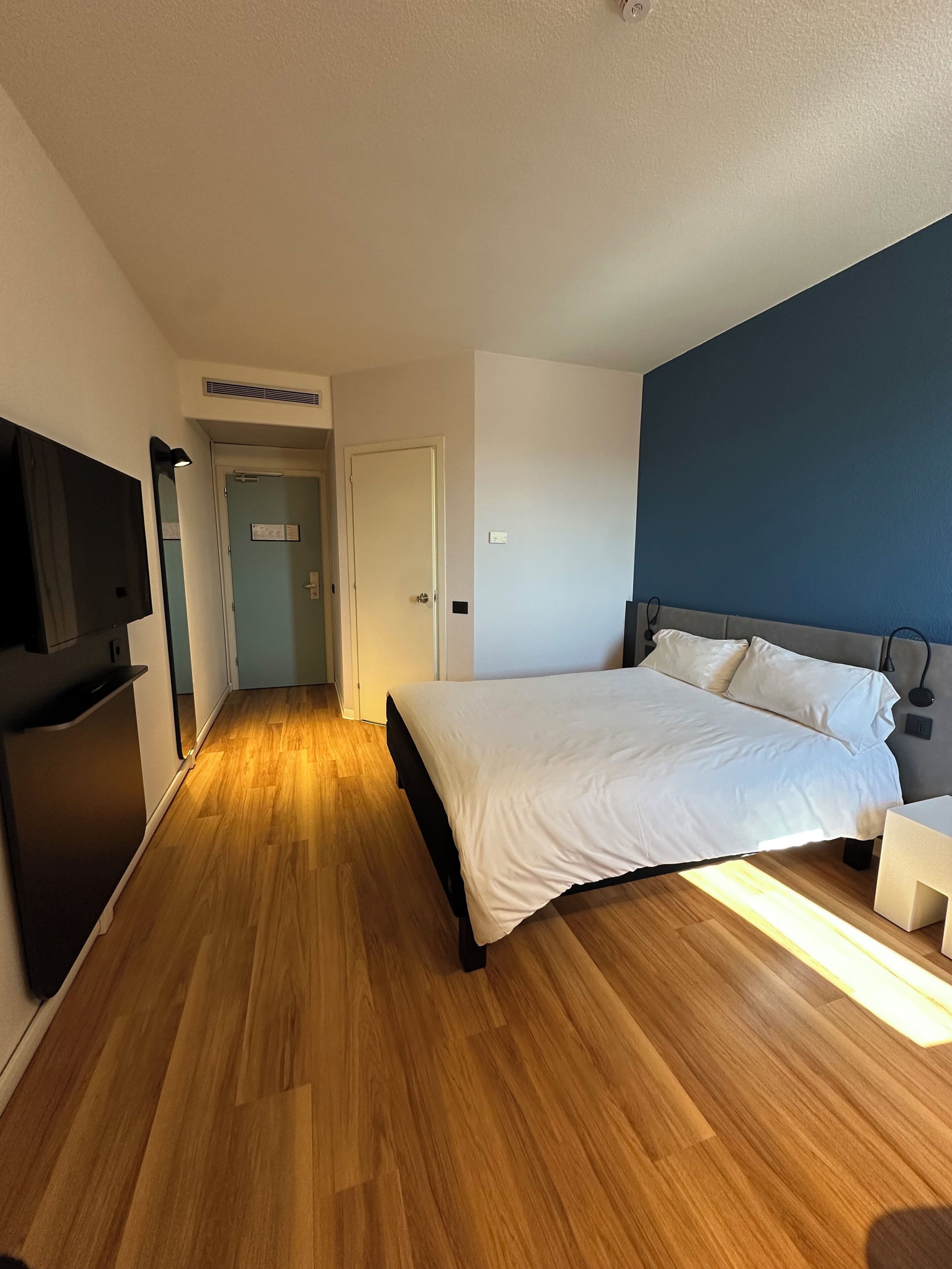 Ein Hotelzimmer mit Holzboden, einem Bett mit weißer Bettwäsche, einem an der Wand befestigten Flachbildfernseher und einer Tür zu einem weiteren Raum.