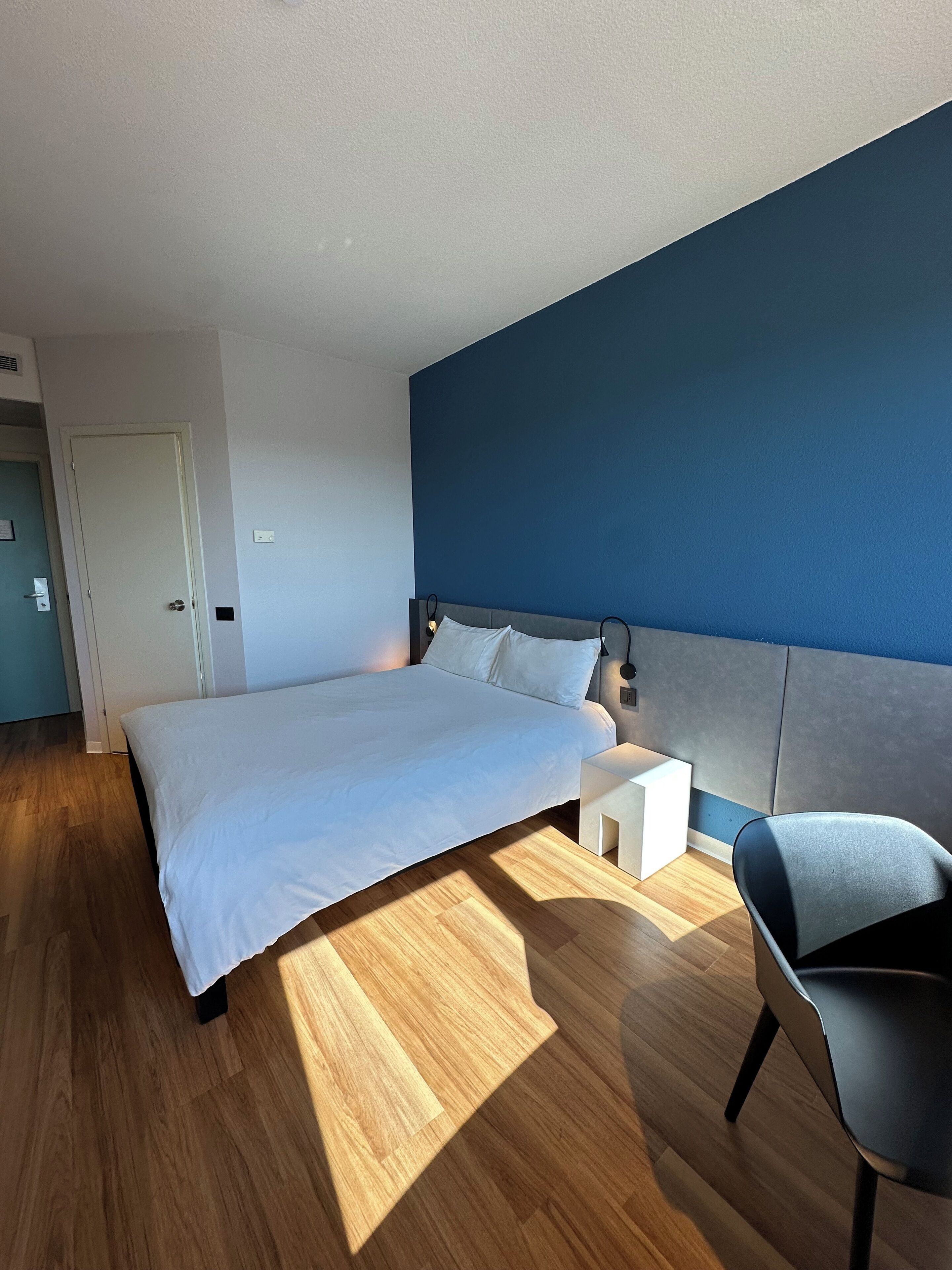 Quarto de hotel moderno com uma cama grande, mesa de cabeceira, uma cadeira e uma parede azul.