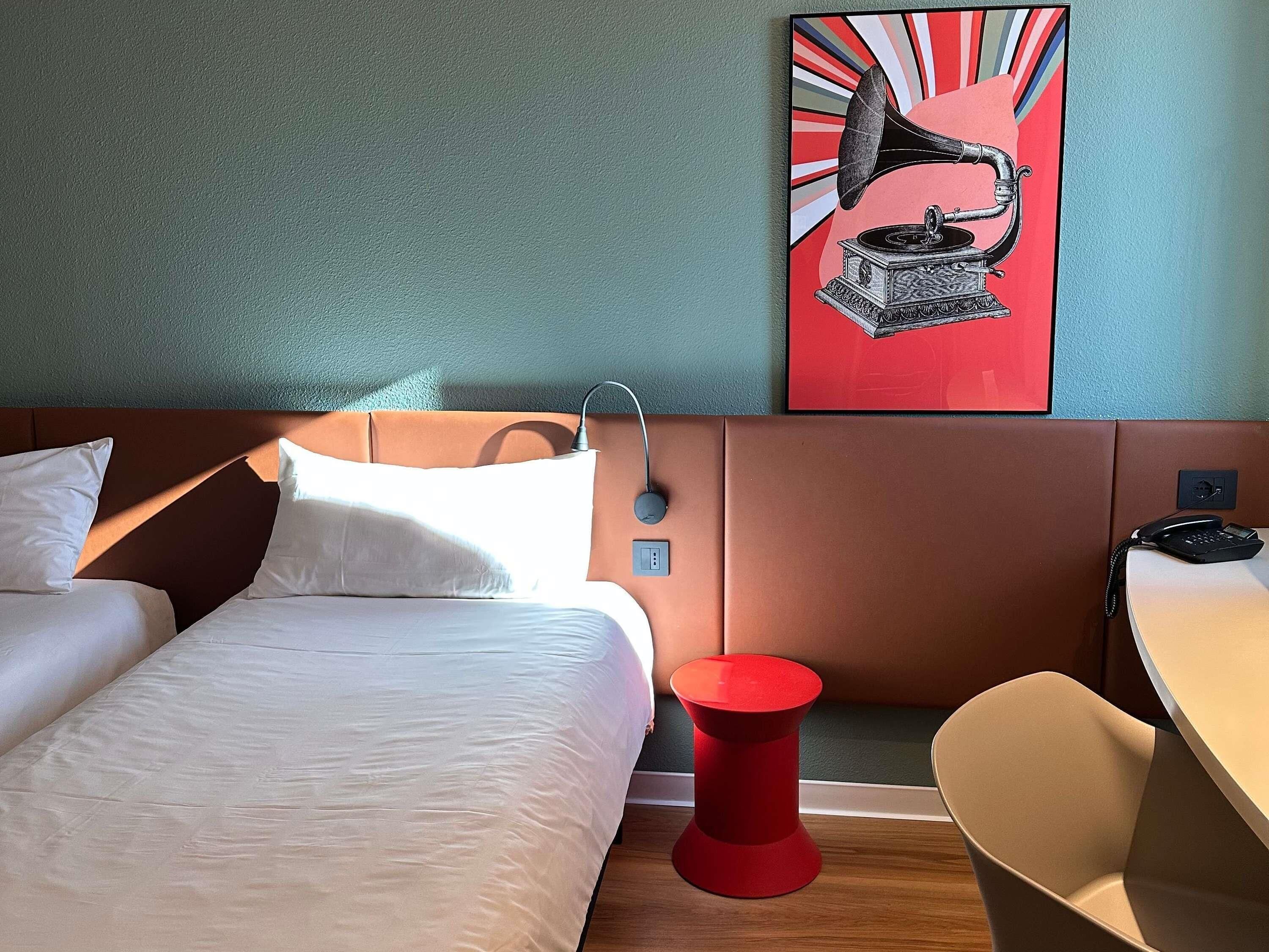Une chambre d’hôtel avec un lit, une table de chevet rouge, une chaise, un téléphone fixé au mur et un tableau représentant un gramophone.