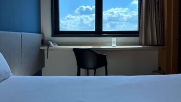 Una habitación de hotel con cama, escritorio con silla, ventana con vista al cielo y nubes, y teléfono sobre el escritorio.