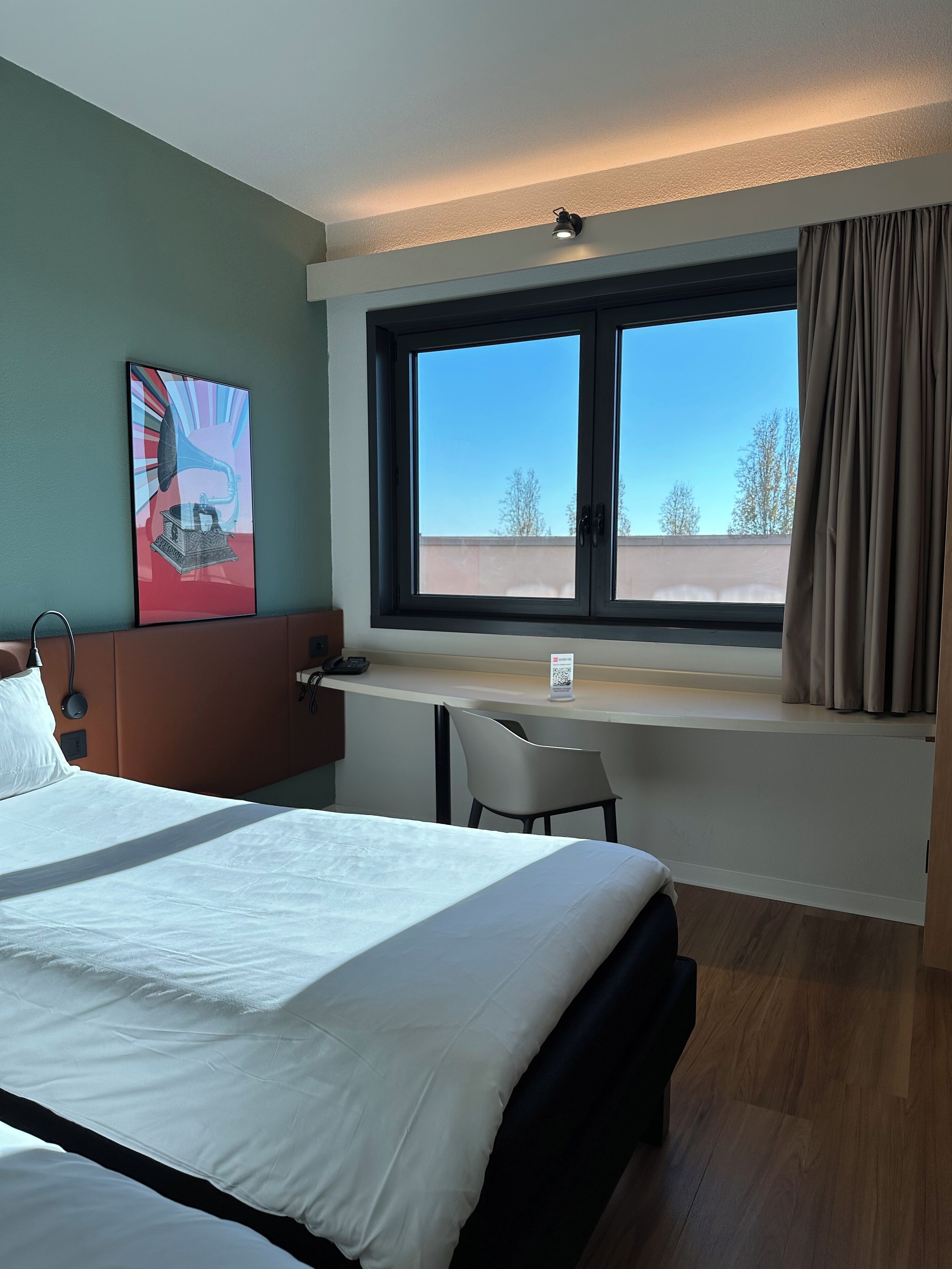 A hotel room with a large bed, a desk with a chair, a window with curtains, and a wall-mounted telephone.