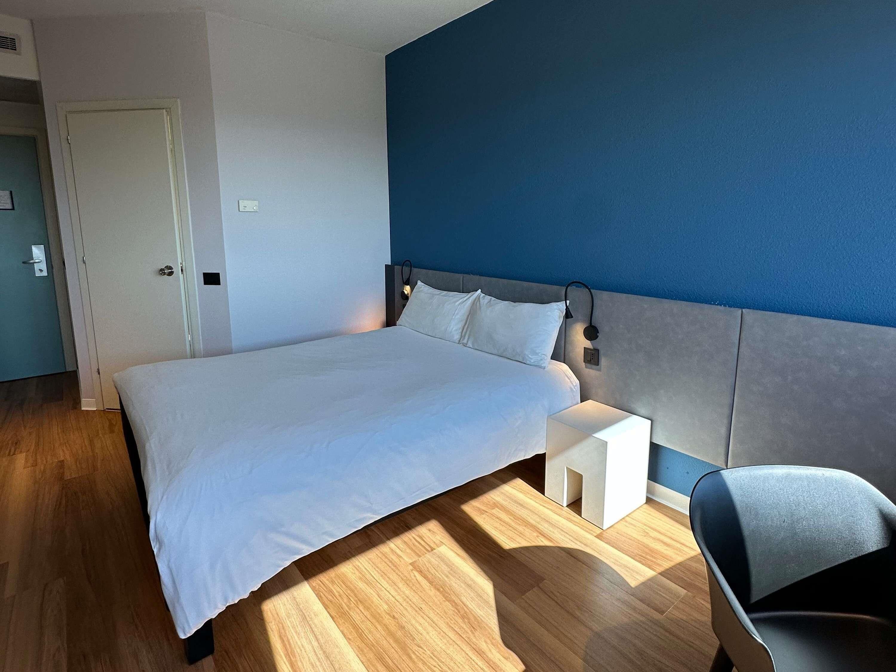 A modern hotel room with a large bed, a bedside table, a chair, and a blue accent wall.