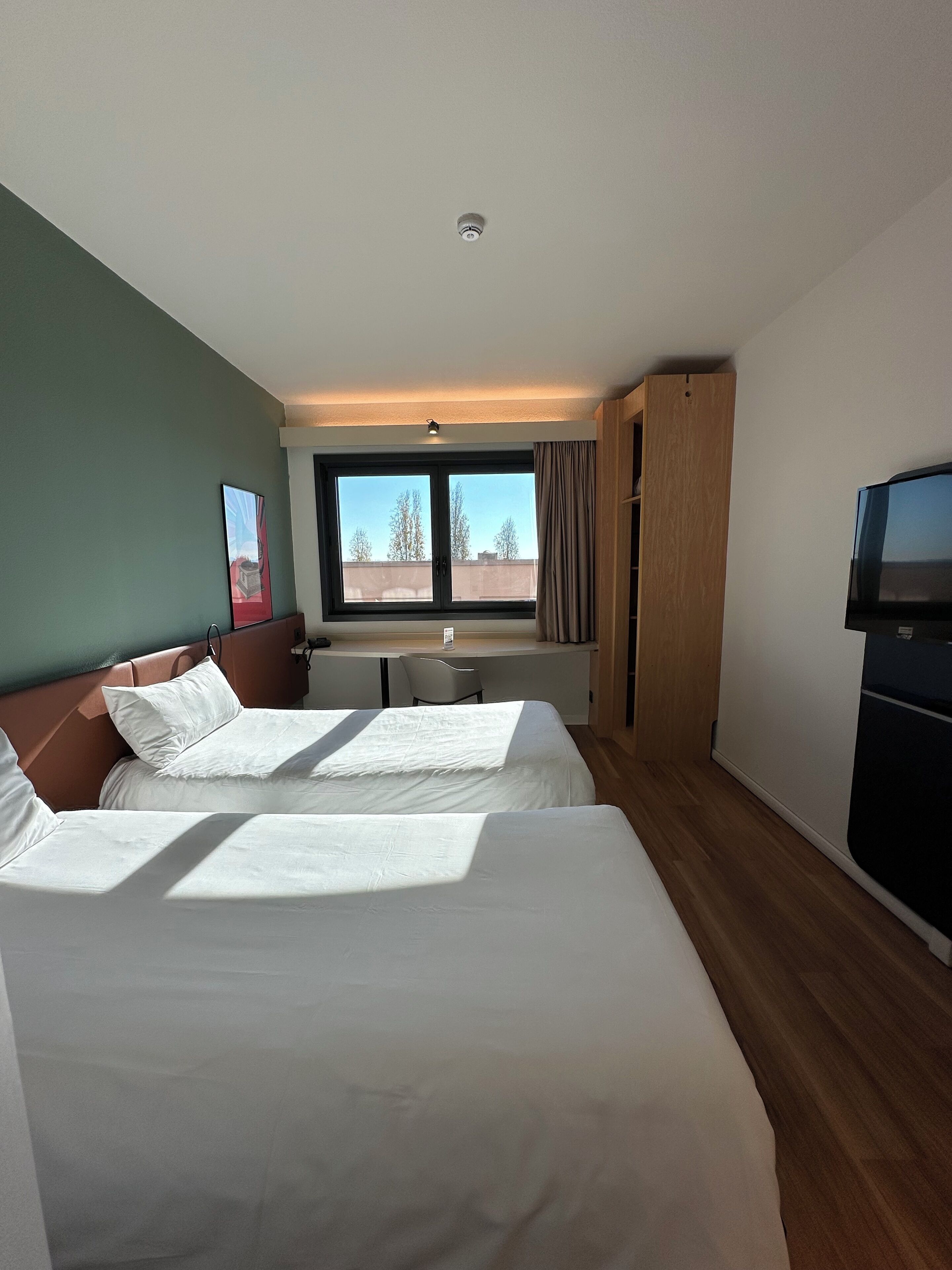Hotellihuone, jossa on kaksi sänkyä, työpöytä, tuoli, televisio ja ikkuna, jossa on verhot.