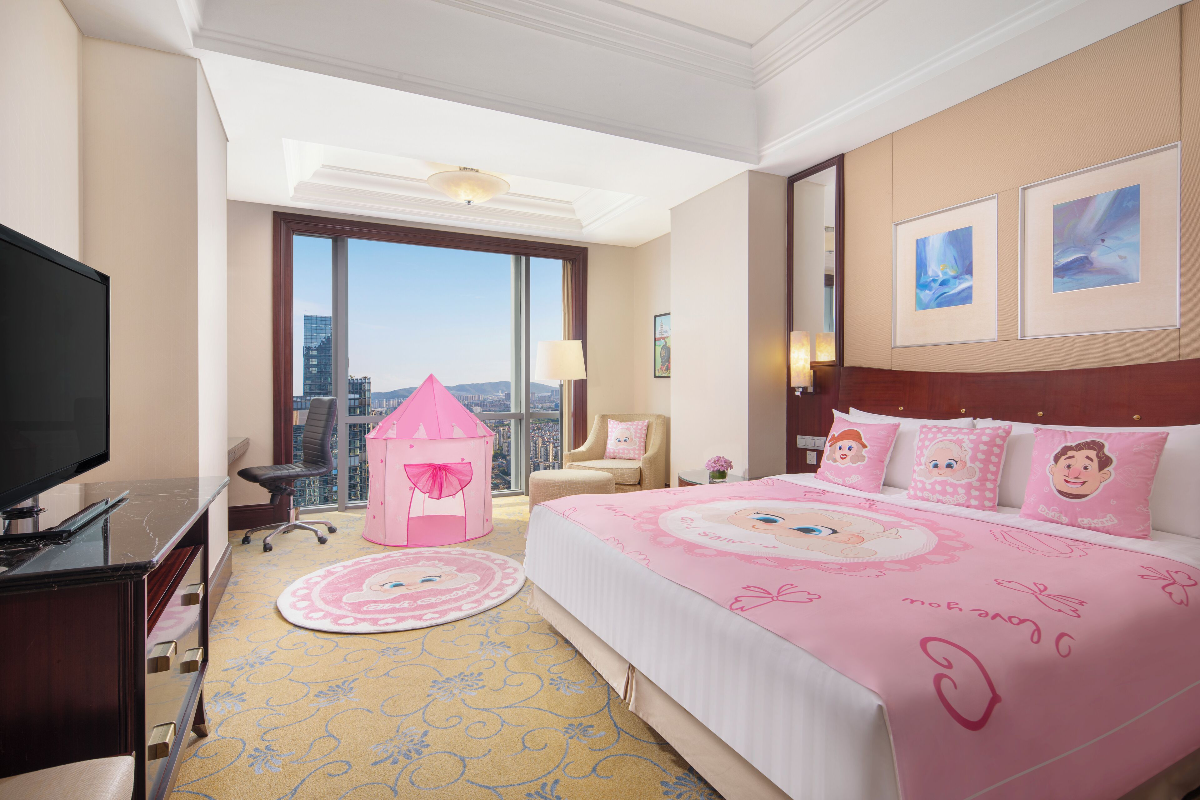 Habitación de hotel con una cama grande, televisión, escritorio, silla, una casita de juegos rosa y vista a la ciudad.