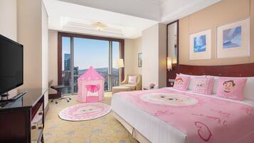 A hotel room with a large bed, a television, a desk, a chair, a pink playhouse, and a view of the city.
