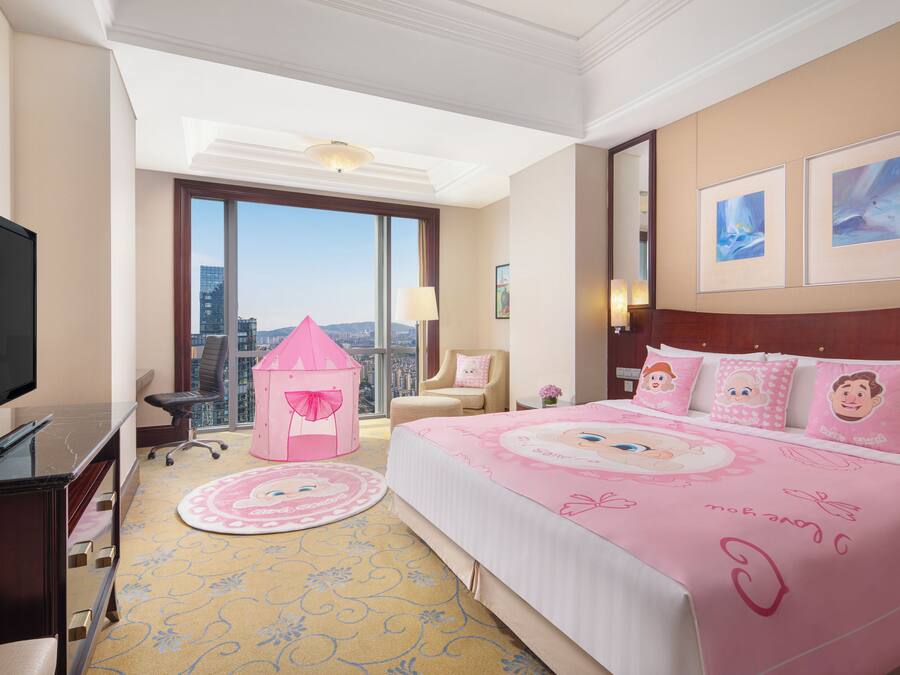 A hotel room with a large bed, a television, a desk, a chair, a pink playhouse, and a view of the city.