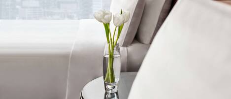 A modern living room with a glass vase containing white flowers on a round table, a city view through large windows, and a sofa with white cushions.