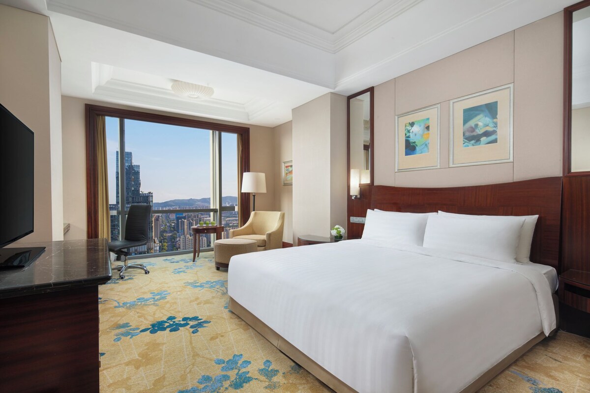 Habitación de hotel con una cama grande, un escritorio con computadora, una silla, una lámpara y vistas a la ciudad.