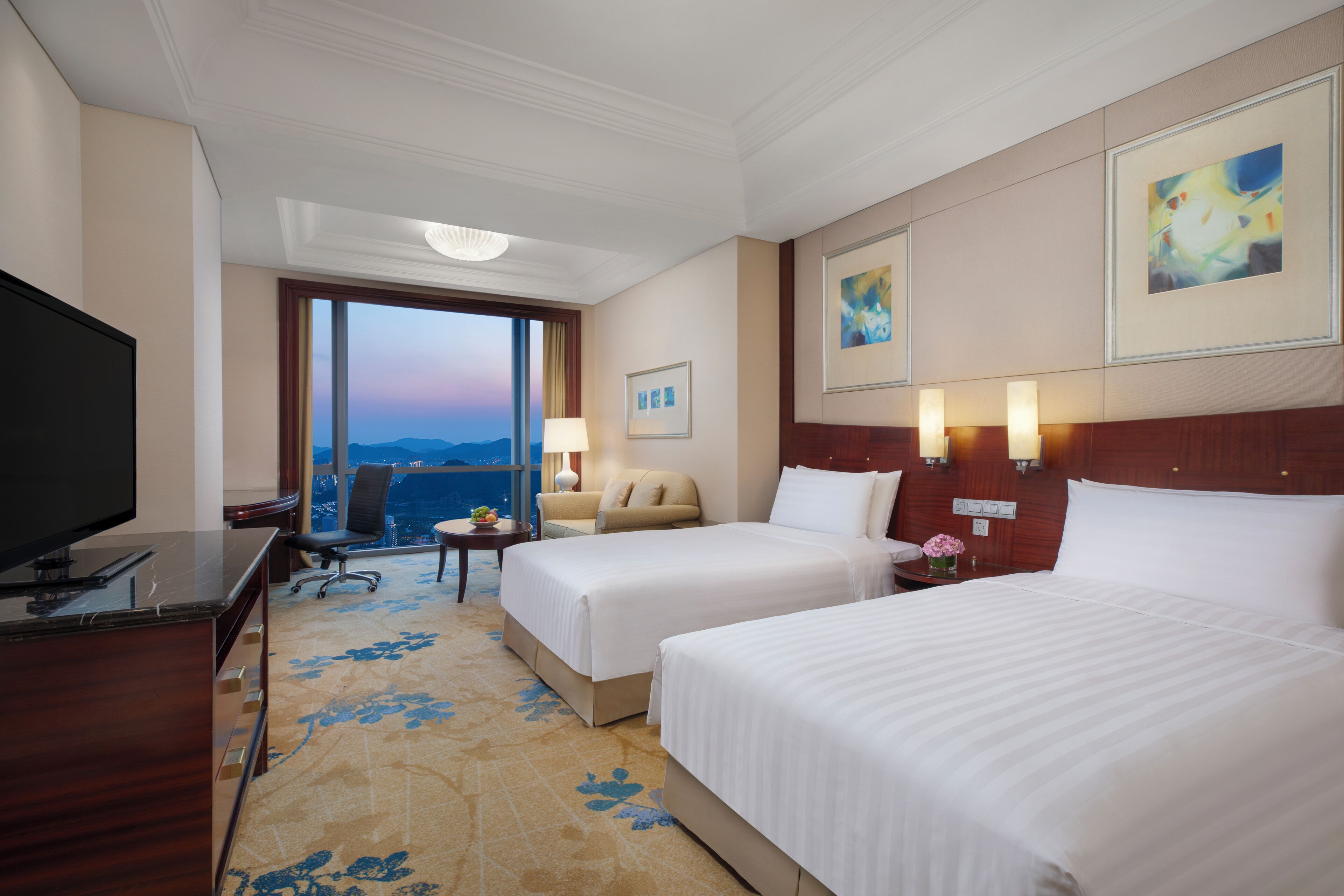 A hotel room with two beds, a desk with a TV, a chair, a lamp, and a view of the mountains.