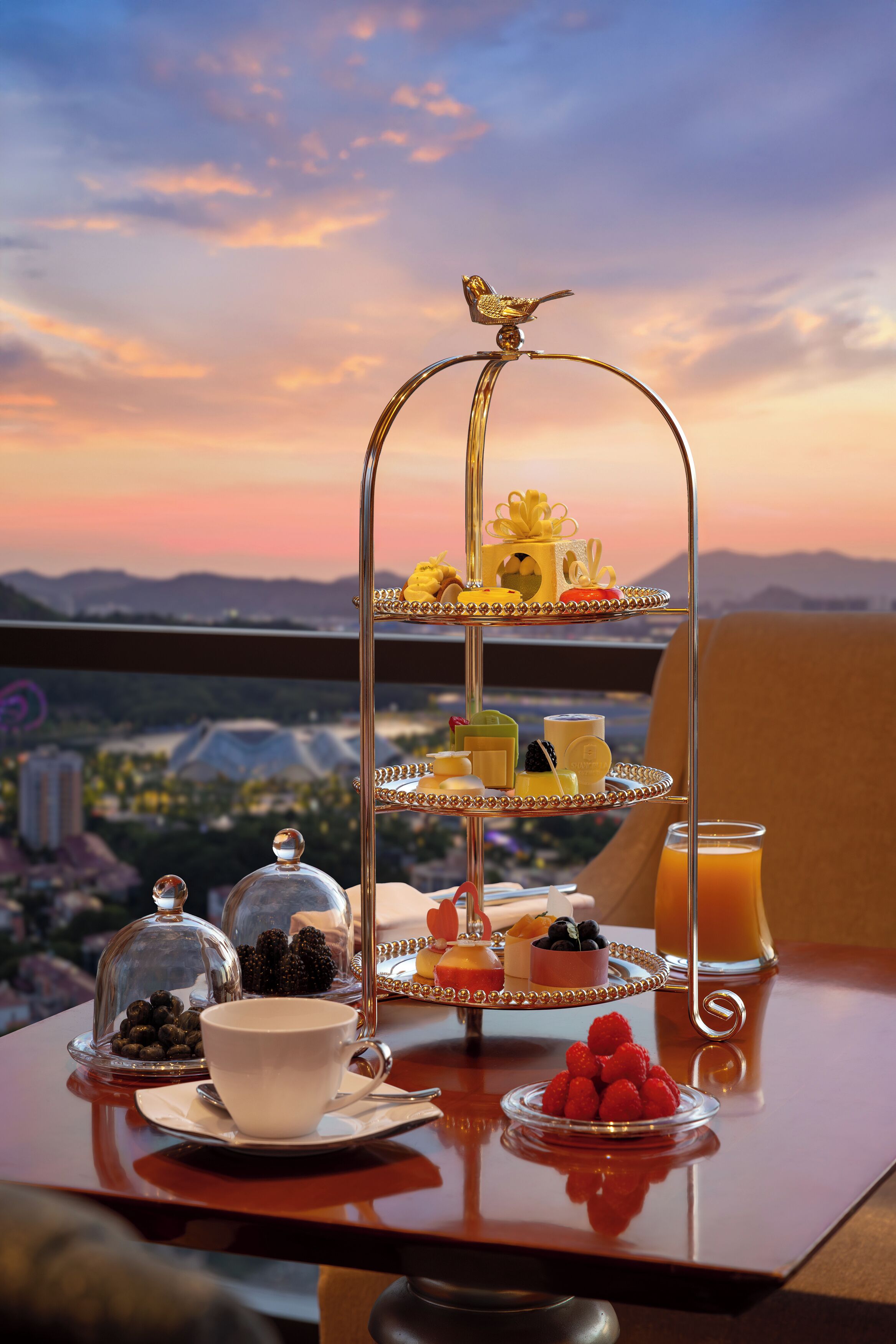 A three-tiered dessert stand with various sweets and fruits, a glass of orange juice, and a cup on a table with a scenic view in the background.