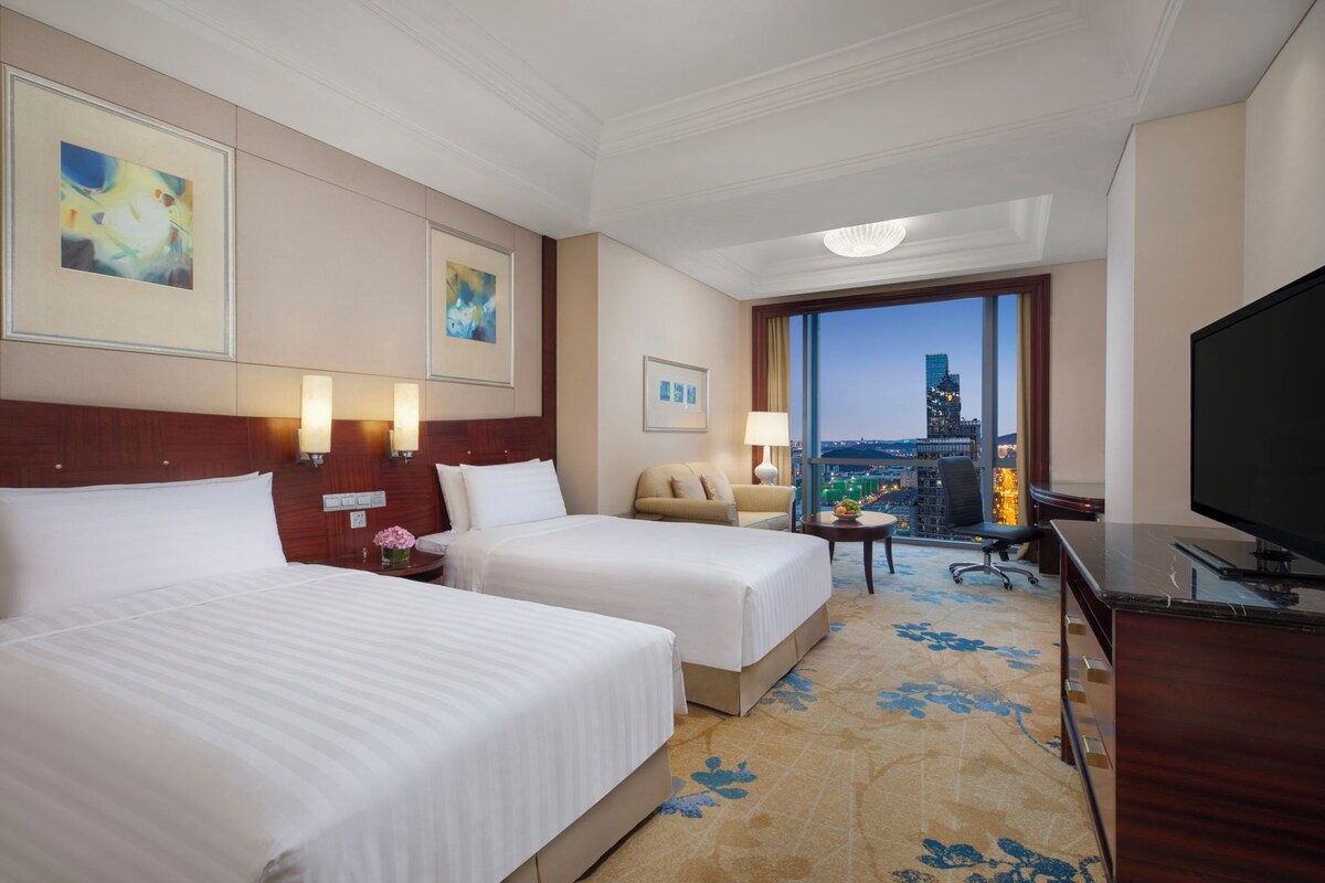 Habitación de hotel con dos camas, un escritorio, un televisor y vista a un paisaje urbano.