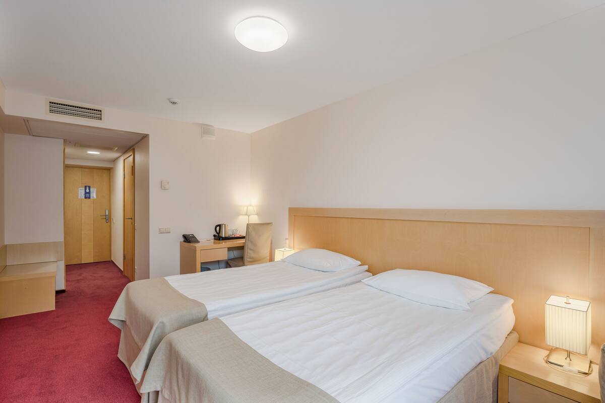 A hotel room with two beds, a desk, a chair, a nightstand with a lamp, and a door leading to another room.