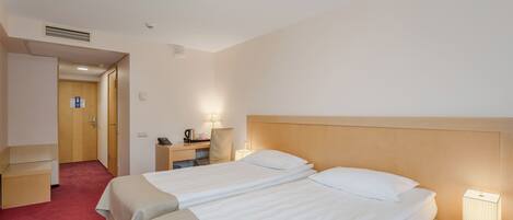 A hotel room with two beds, a desk, a chair, a nightstand with a lamp, and a door leading to another room.
