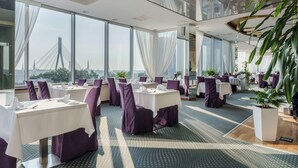 A modern dining area with tables set for service, large windows offering a view of a bridge, and indoor plants. - Riga Islande Hotel with FREE parking (Riga)