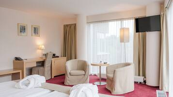 A hotel room with a bed, two armchairs, a desk, a TV, and a window with curtains.