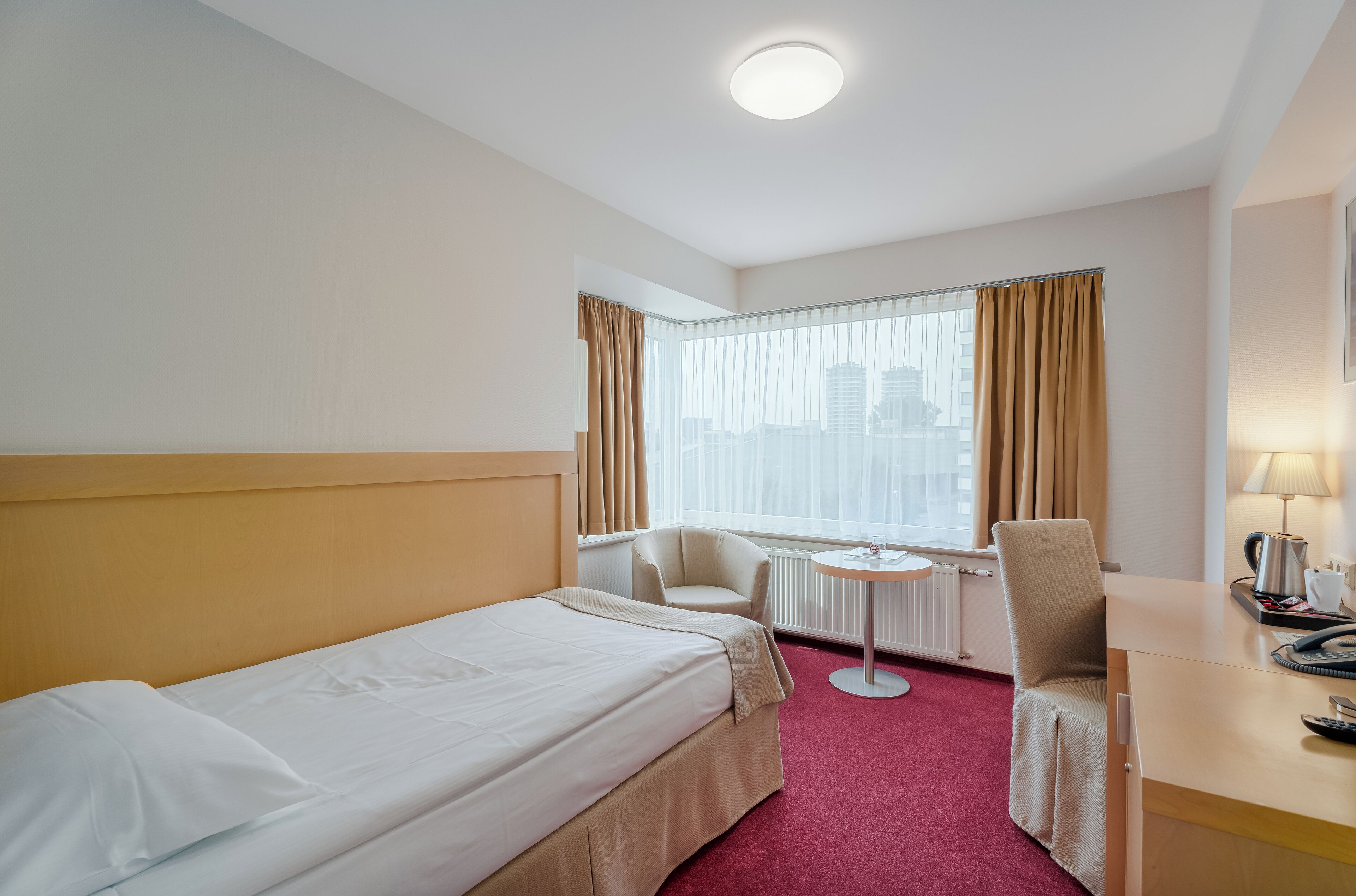 A hotel room with a bed, a desk, a chair, a small table, and a window with curtains.