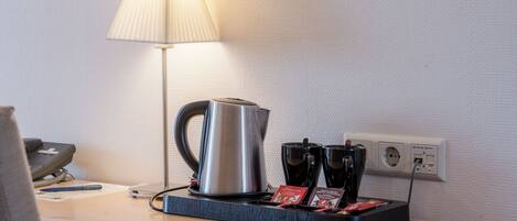 A hotel room with a bedside table featuring a kettle, two cups, a lamp, and a phone.