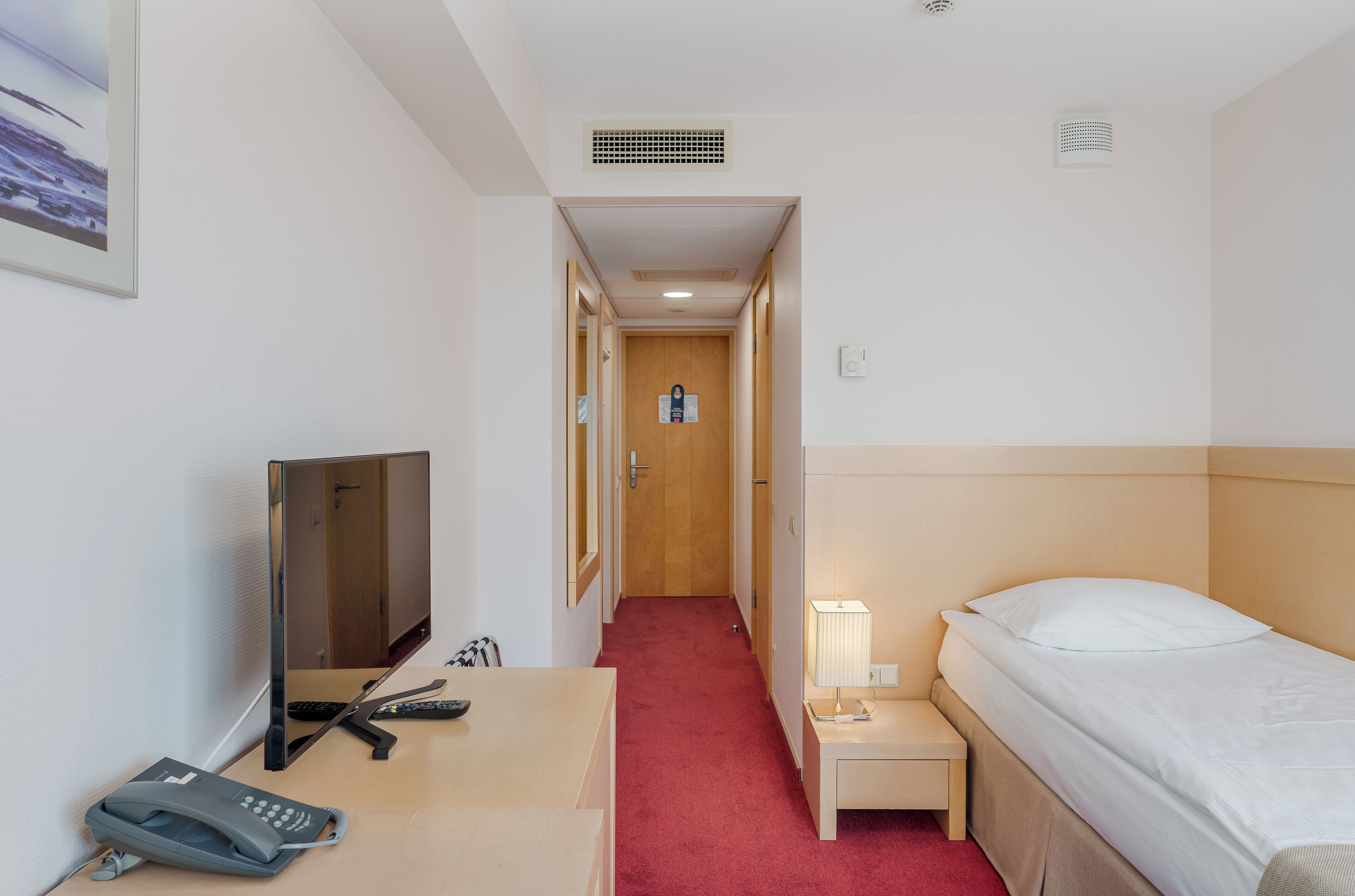 A hotel room with a bed, bedside table, lamp, TV, and a desk with a phone and remote control.