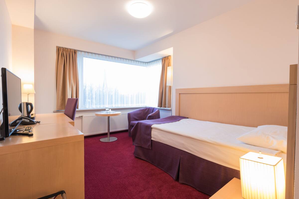 A hotel room with a bed, a desk, a chair, a window with curtains, and a lamp.