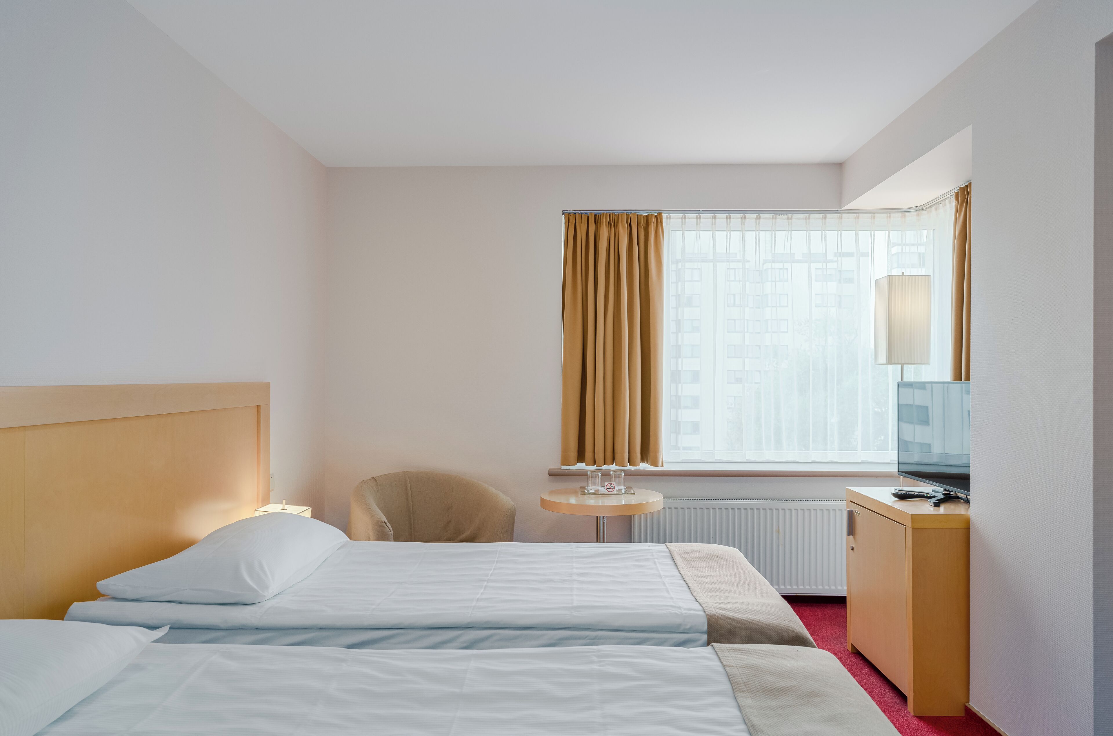 Hotellihuone, jossa on kaksi sänkyä, työpöytä, tuoli, televisio ja ikkuna, jossa on verhot.
