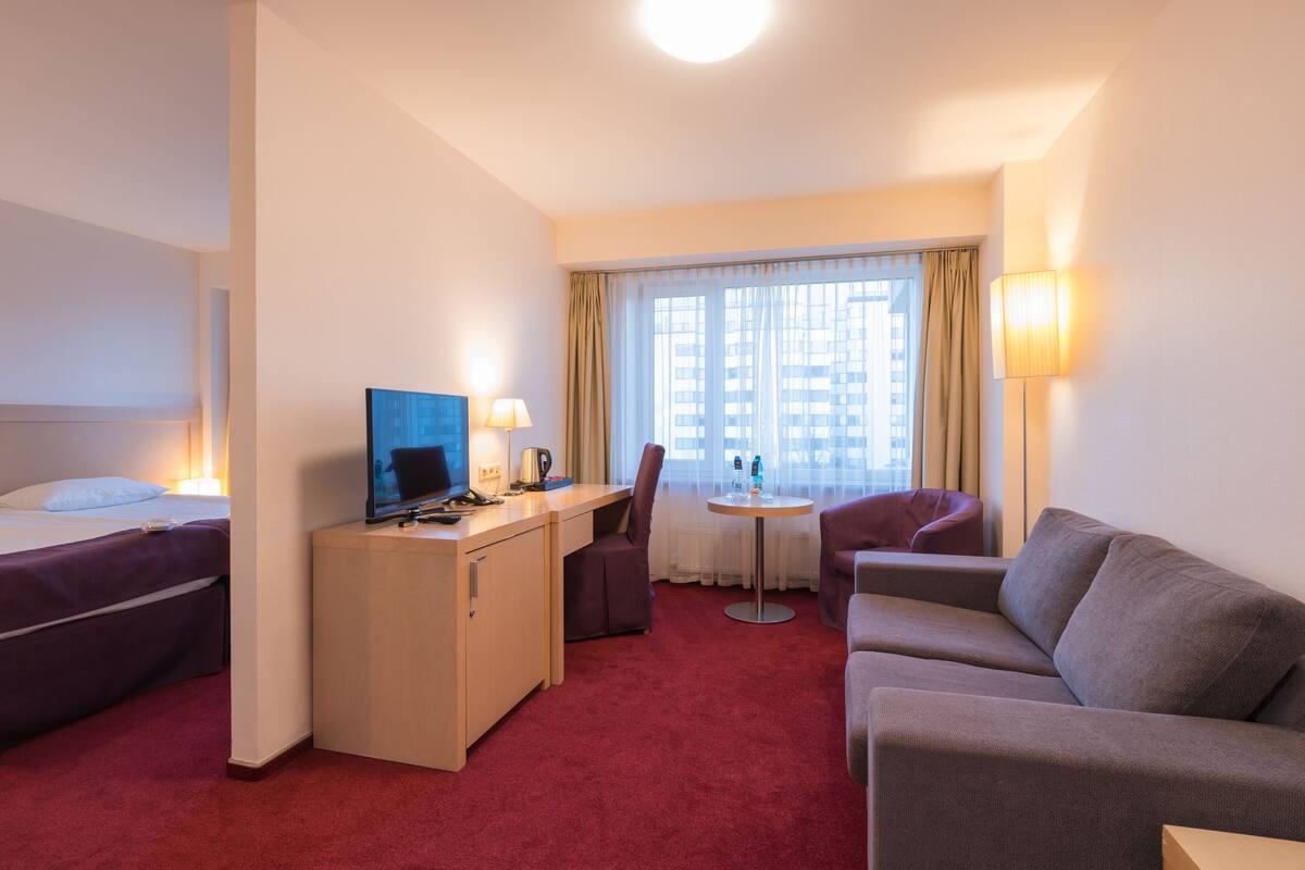 A hotel room with a bed, a desk with a computer, a sofa, and a small table with a lamp.