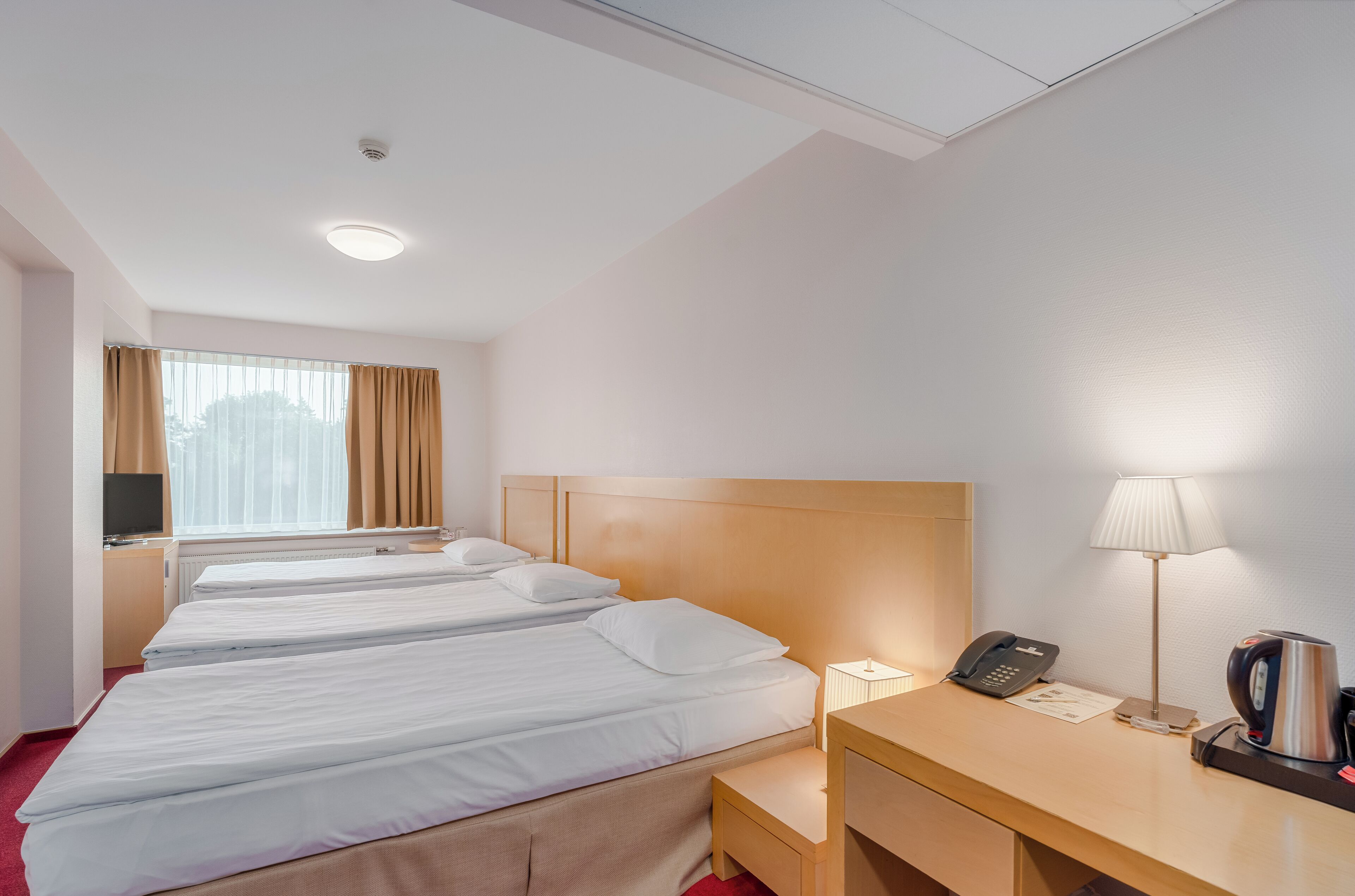 A hotel room with three single beds, a desk with a lamp, a telephone, and a coffee maker.