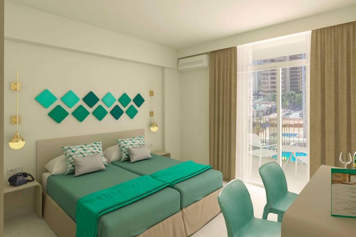 A hotel room with a bed, two bedside tables, a desk with a chair, a window with a view of buildings and a pool, and decorative wall art.